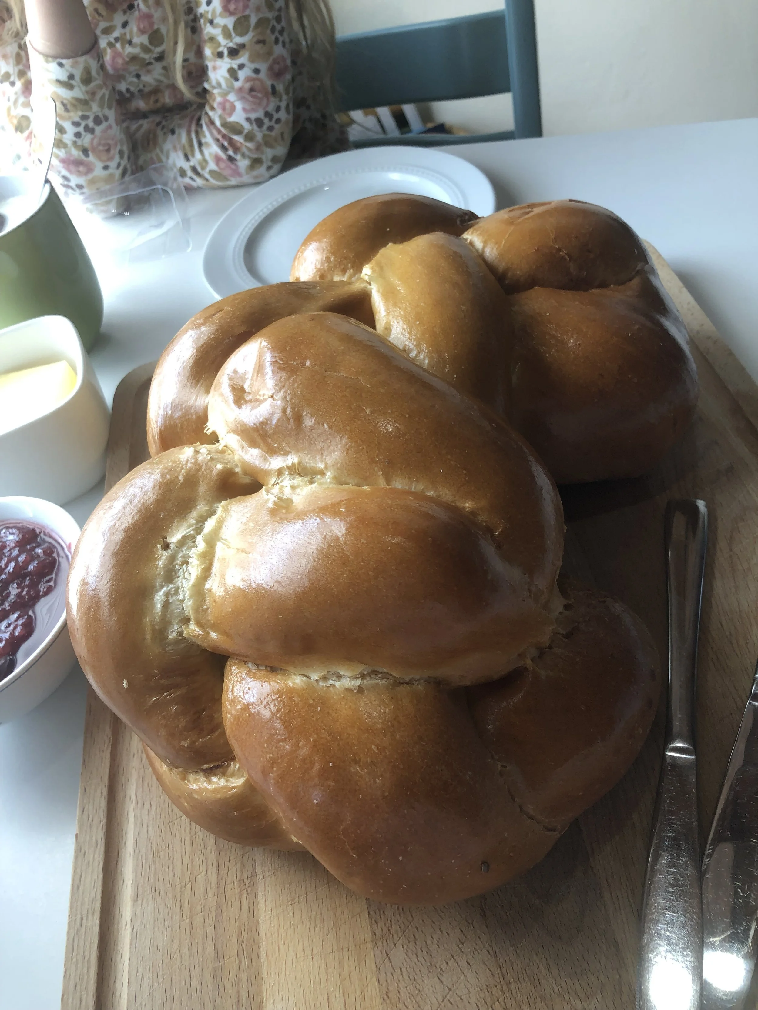 Child (you can only see body) in flower t-shirt sitting at a table with a gigantic braided bread, you can see butter and jam near by - foto by Maja Sofie Kristiansen