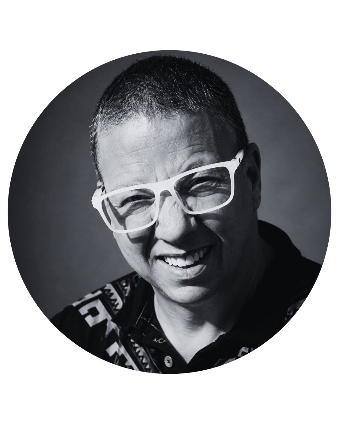 Black and white photo. Paulie is an older man with short-trimmed hair and white-rimmed glasses. He wears a black button-up with an Indigenous pattern along the shoulders.