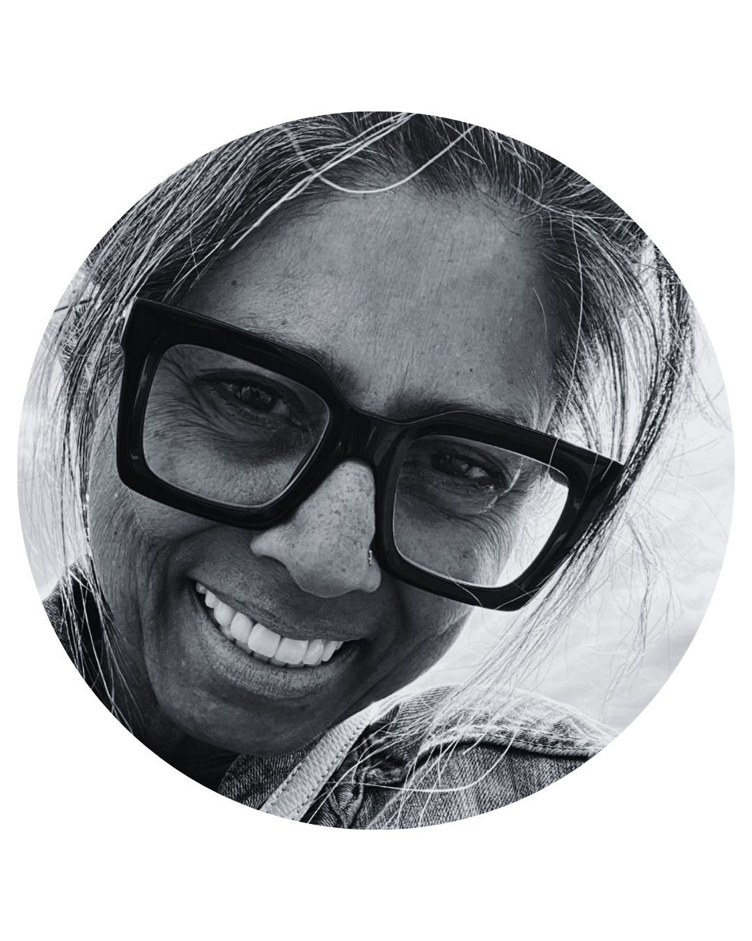 Black and white photo. Salma is an older woman with long salt and pepper hair loosely framing her face. She wears thick black-rimmed square glasses and smiles widely.