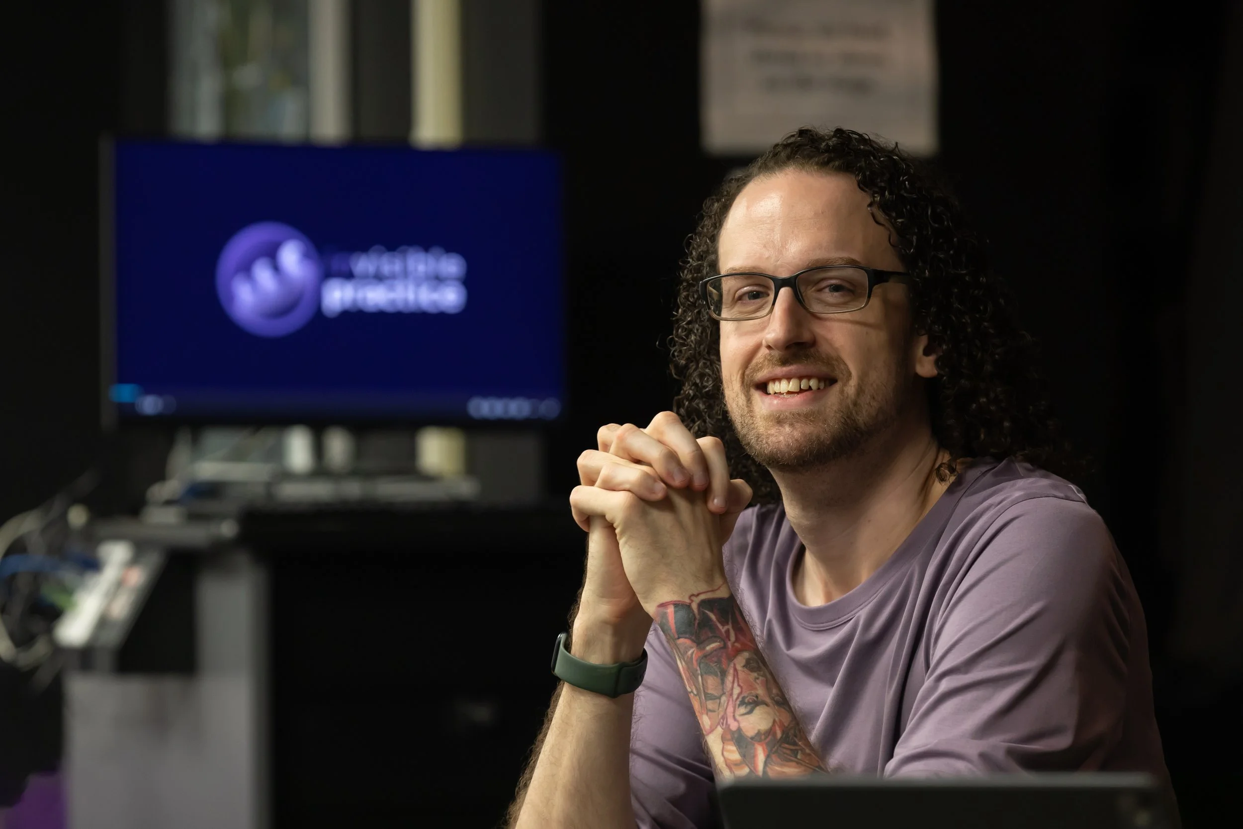 Jan is a young white man with almost shoulder-length curly hair. He sits at a table with his hands clasped near his chin, and gazes at the camera with a relaxed gladness.