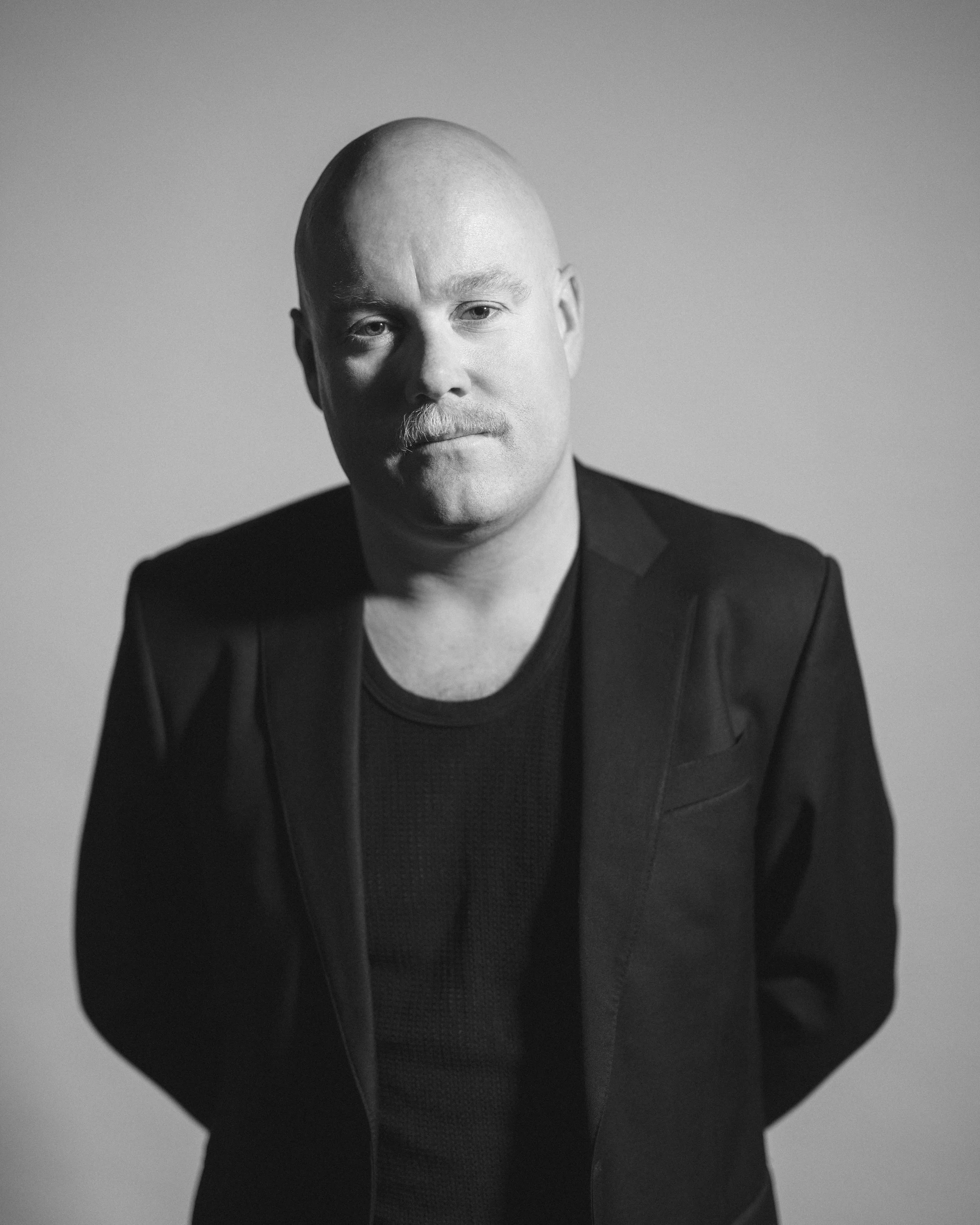 Trent is a bald white man with a mustache wearing all black. He is looking intently at the camera with arms crossed behind his back. He has a hard shadow against the side of his face.