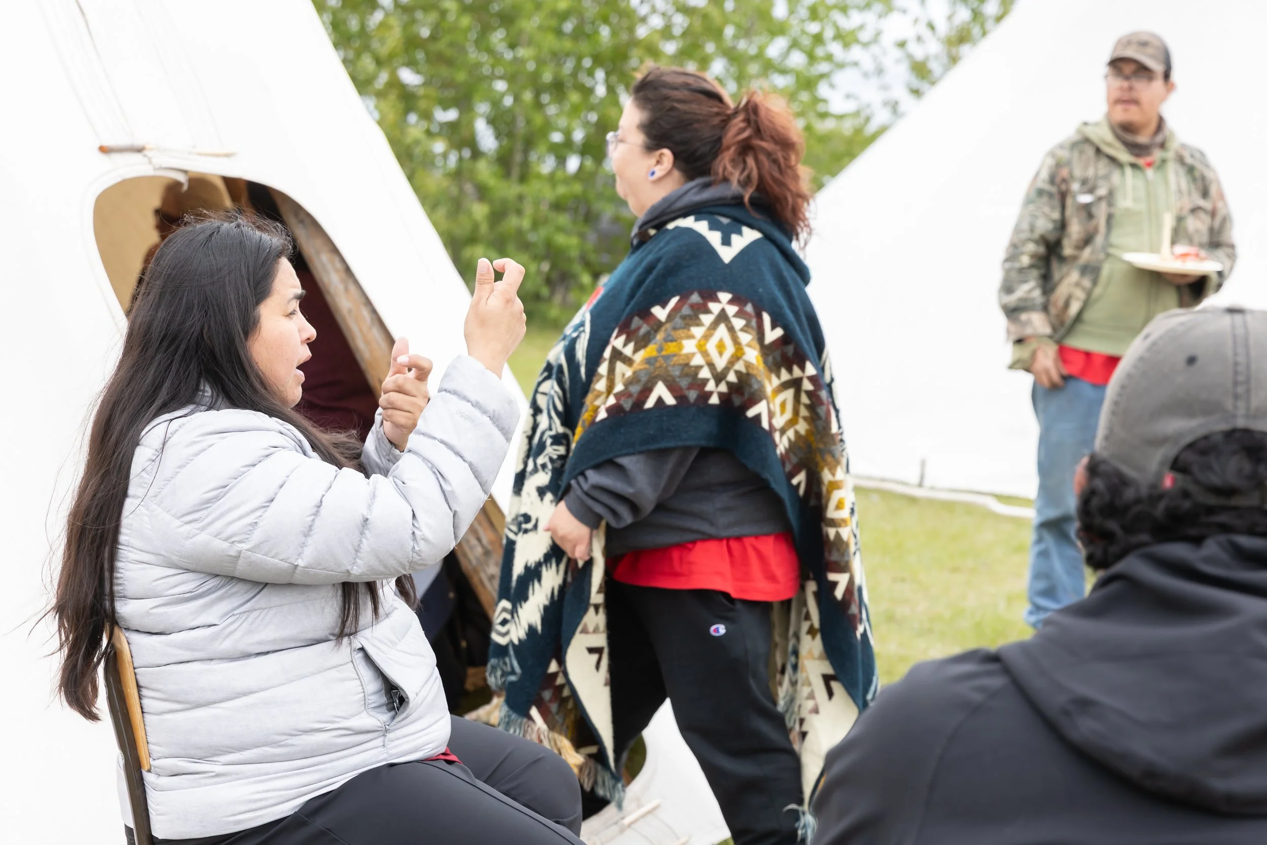 Some folks sit and other stand among two white teepees.