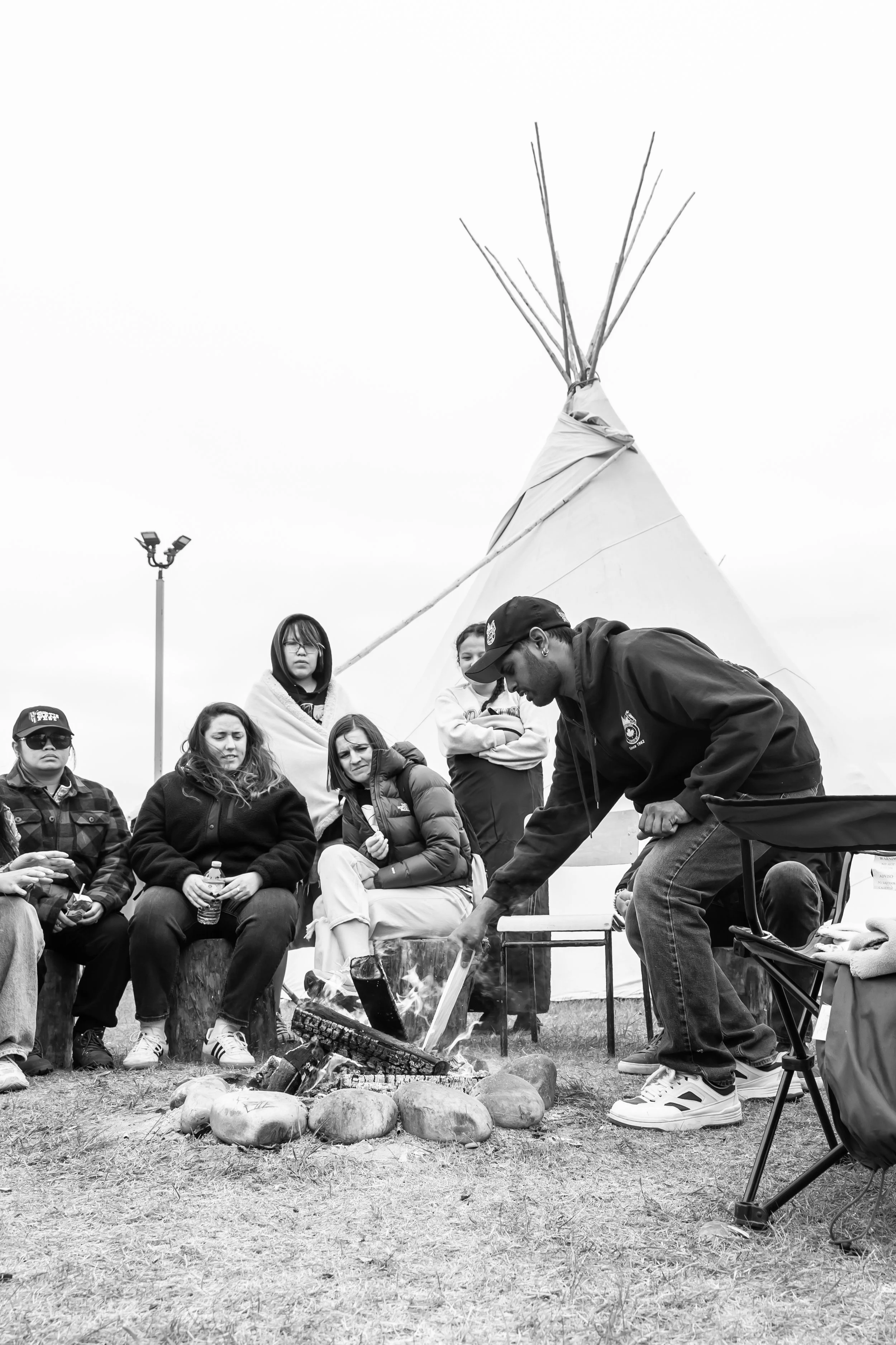 Black and white. Several people gather around a campfire with a teepee in the background.