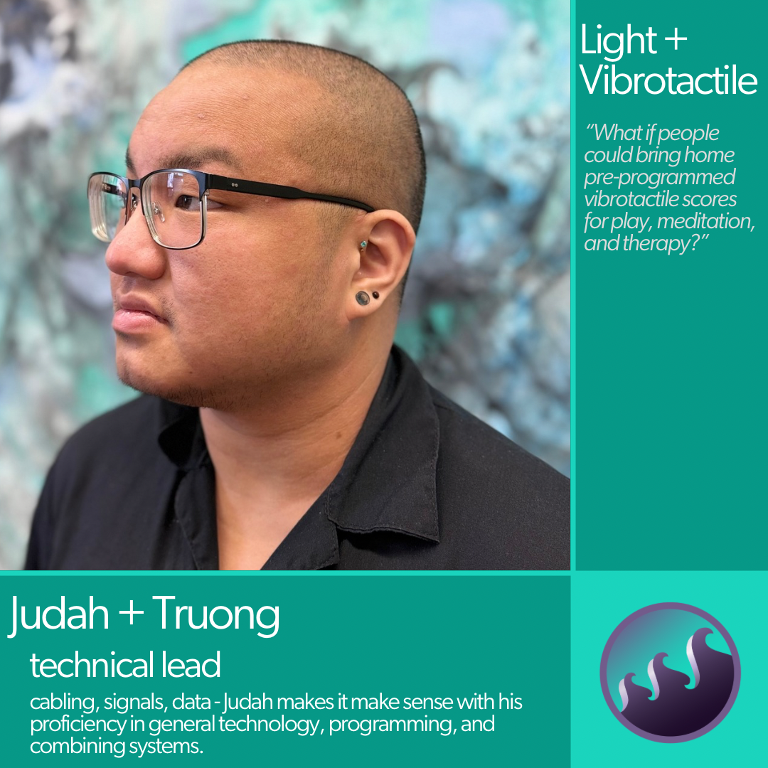 Judah is a young Asian person with buzzed hair, glasses, and several piercings. He wears a black open-neck shirt and looks up and to the left while situated against a swirly blue-teal painting.