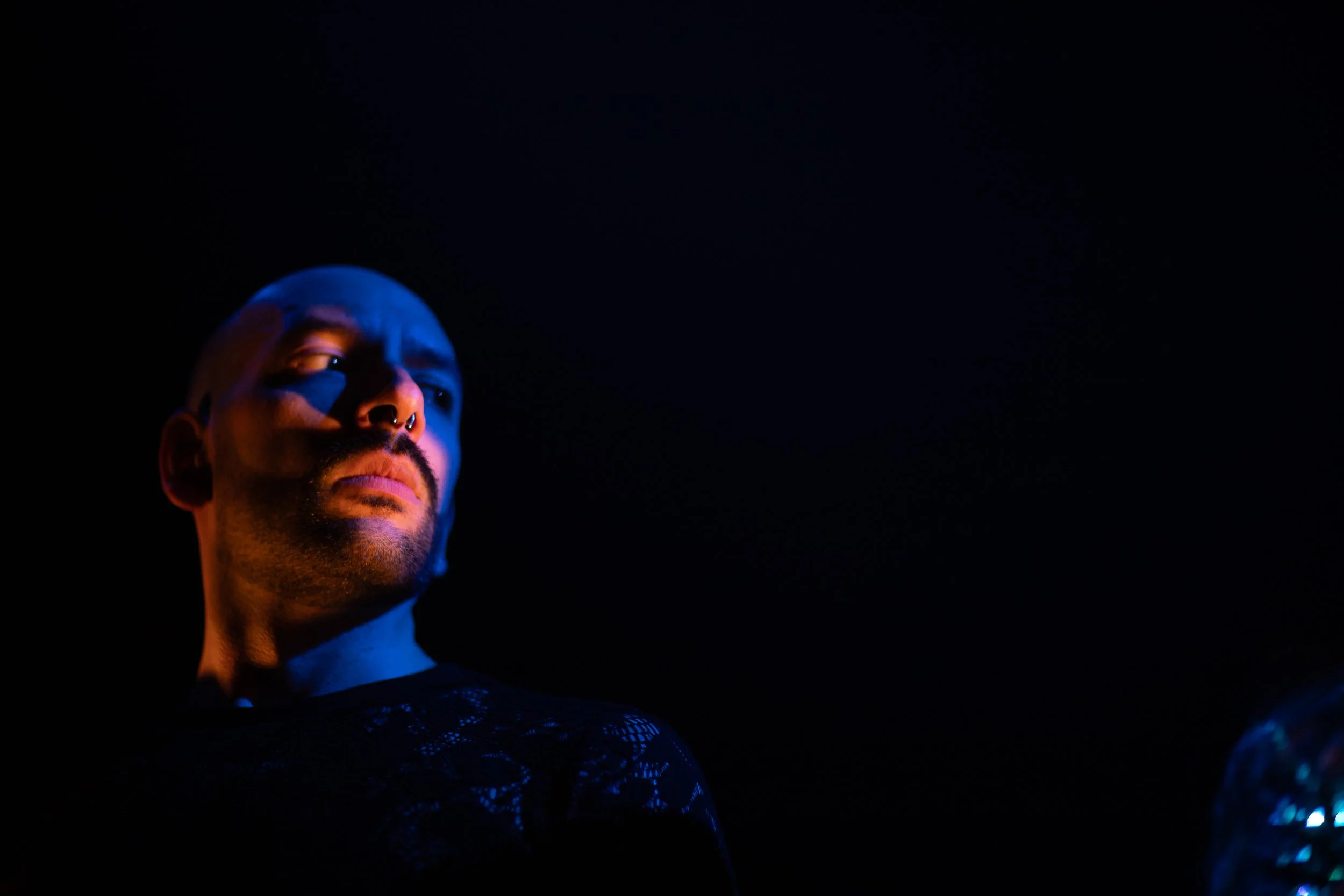 Connor’s face is starkly lit from the sides in a dark space, in tones of blue and light pink. He is looking to one side at an angle with a solemn expression. He is a young white man with neatly trimmed beard and a small silver bull nose ring.