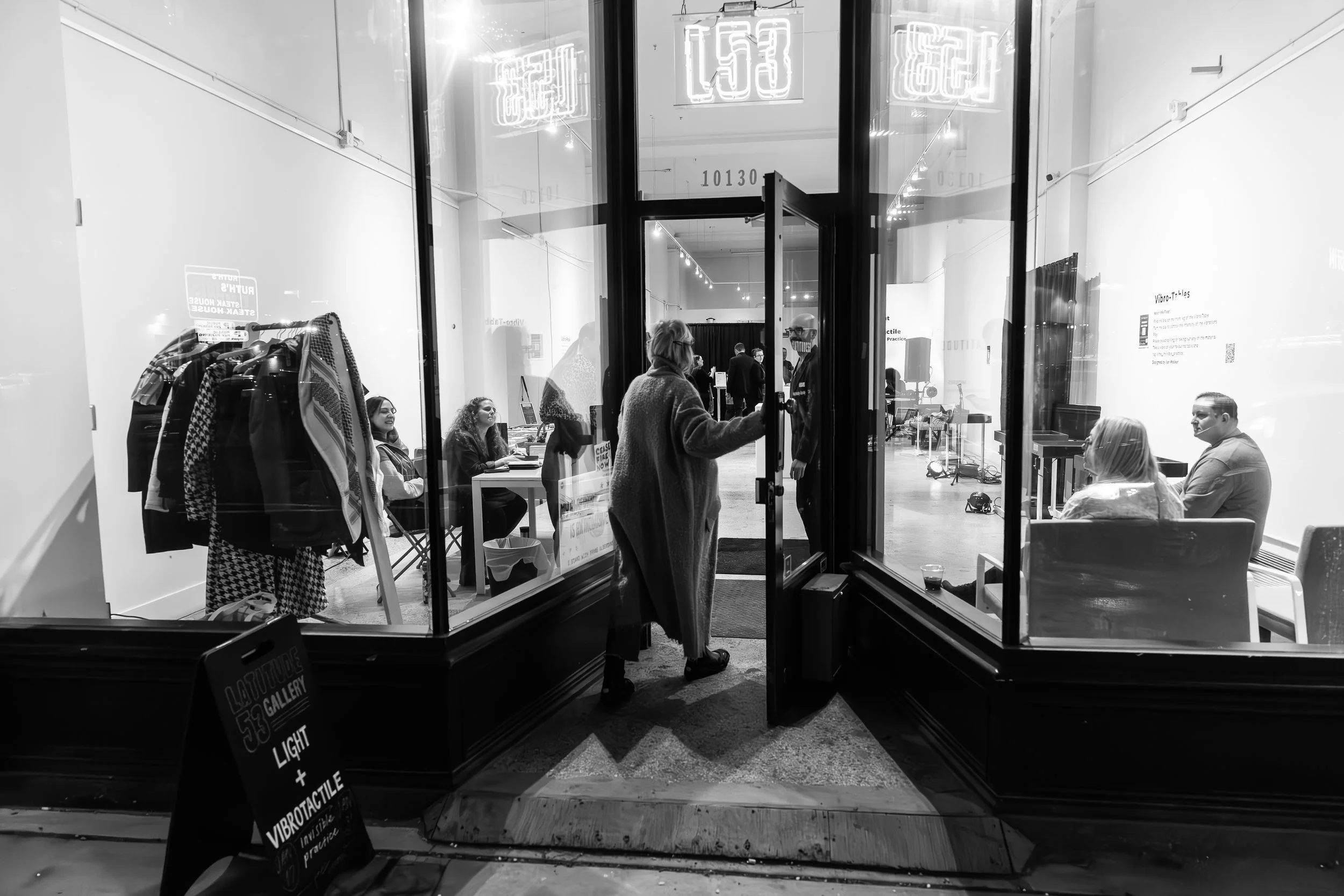 Black and white photo of a person entering through a glass door, flanked by large glass bay windows showing an art gallery space.  It is glowingly lit and already filled with people. A neon sign hangs above - “L53.”