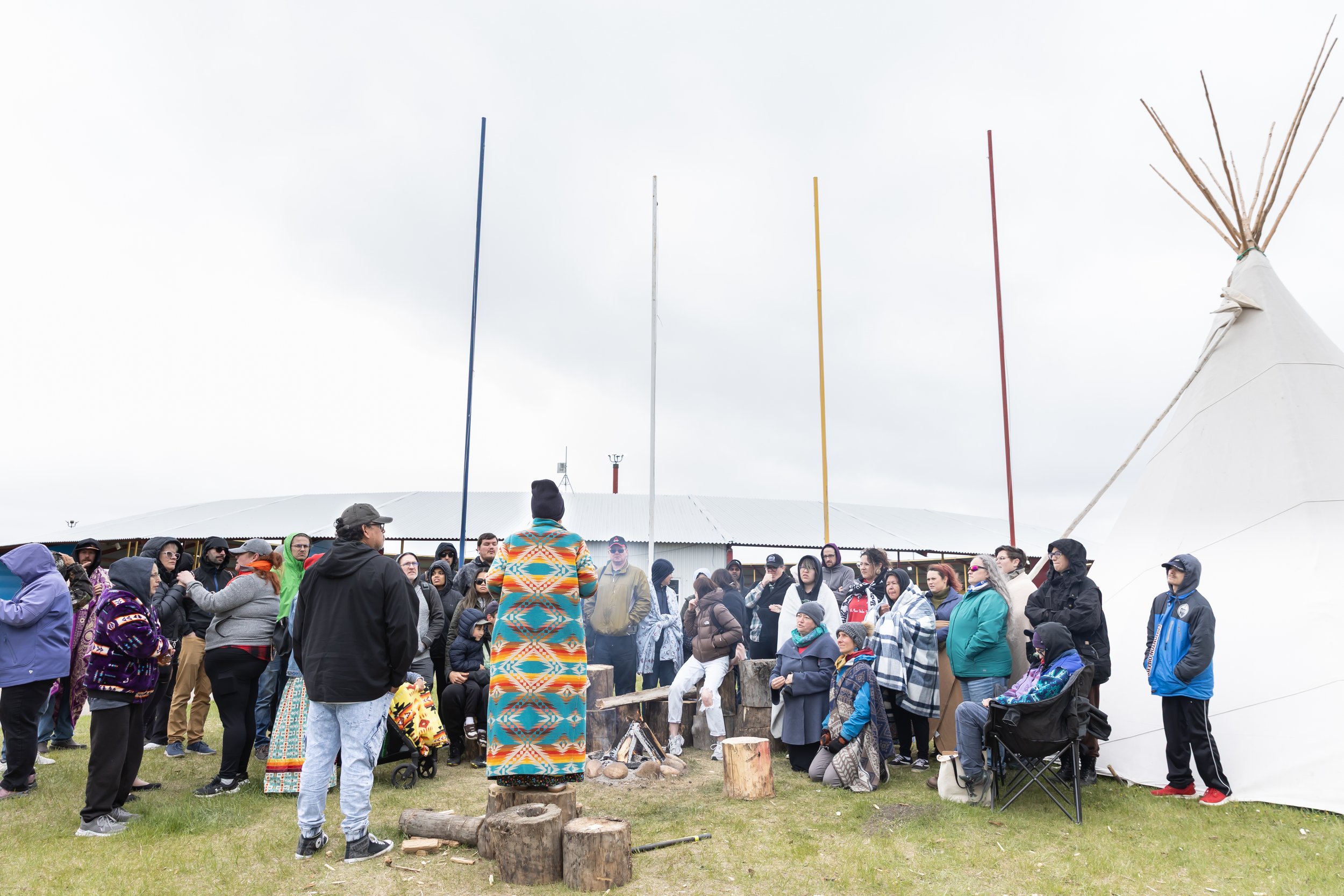 A further-away shot of the gathered crowd next to a white teepee. Two people address them with their backs to the camera. One of them stands on a stump wearing a bright Indigenous patterned jacket.