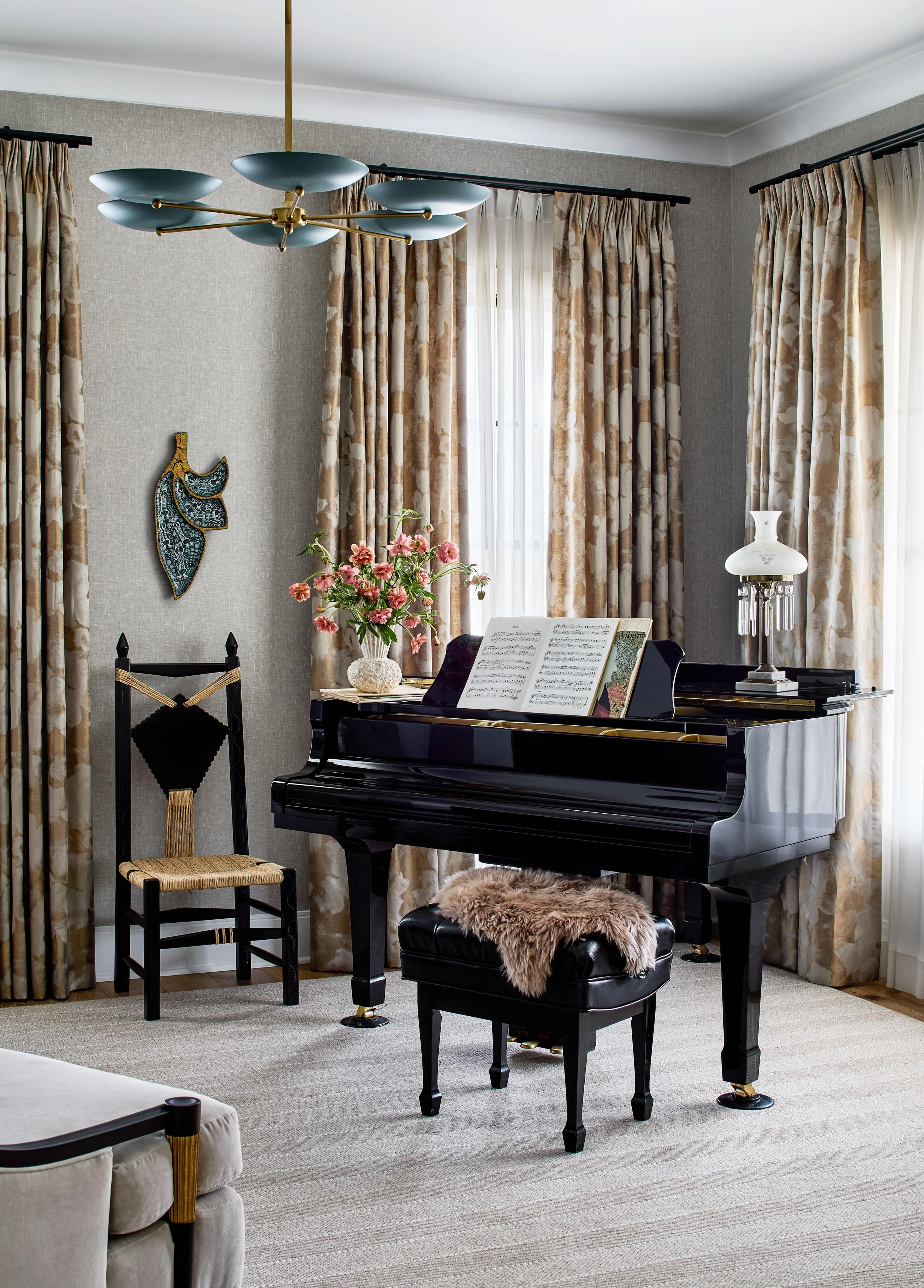 Living room with a black grand piano, a furry bench, a wooden chair, floral curtains, a vase with pink flowers, and a modern chandelier.
