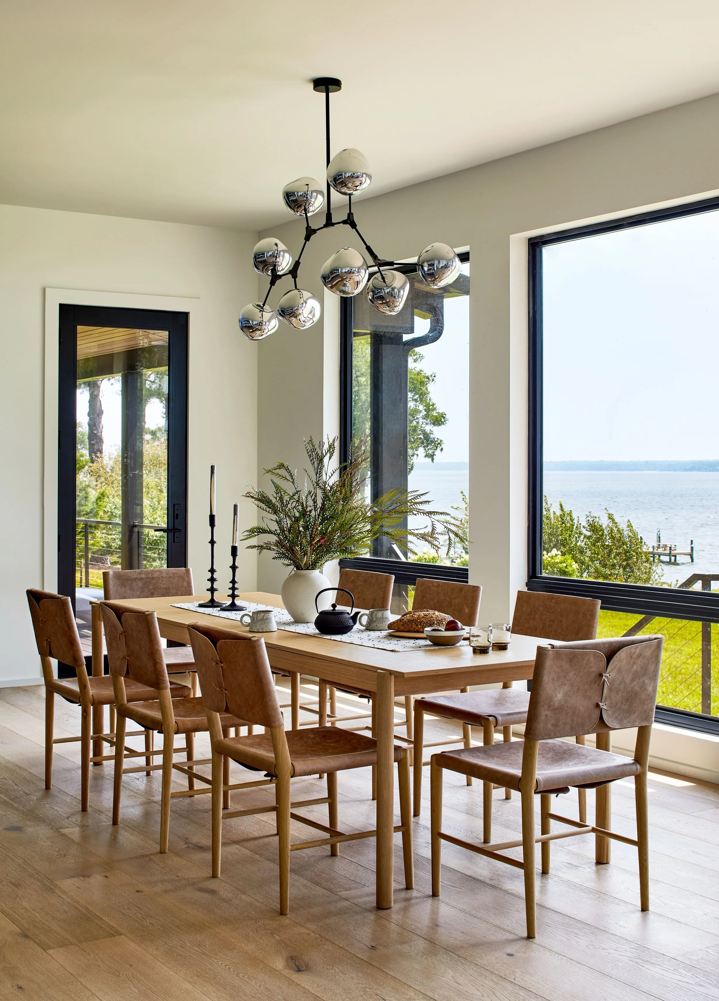 Dining room with a wooden dining table, ten chairs, large window overlooking water and trees, modern chandelier, vase with greenery, and breakfast items.