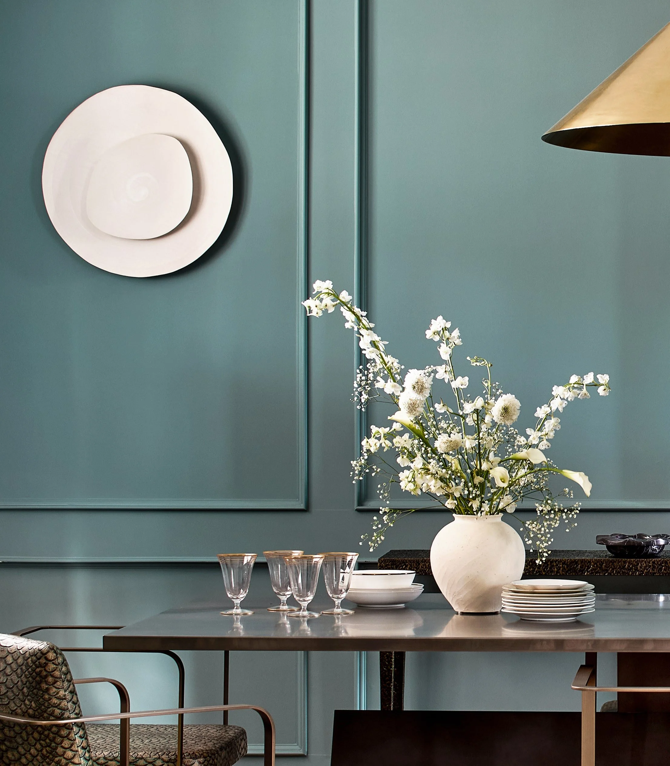 A dining room table with a white vase filled with white flowers, surrounded by empty glasses and plates, against a teal paneled wall decorated with circular white wall art.