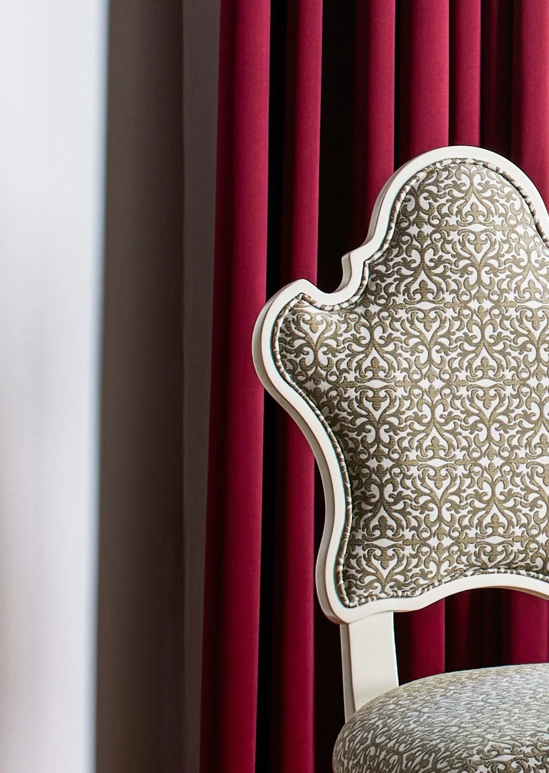 Close-up of an ornate chair with a beige and brown patterned fabric, featuring a white carved frame, positioned in front of a set of red velvet curtains.