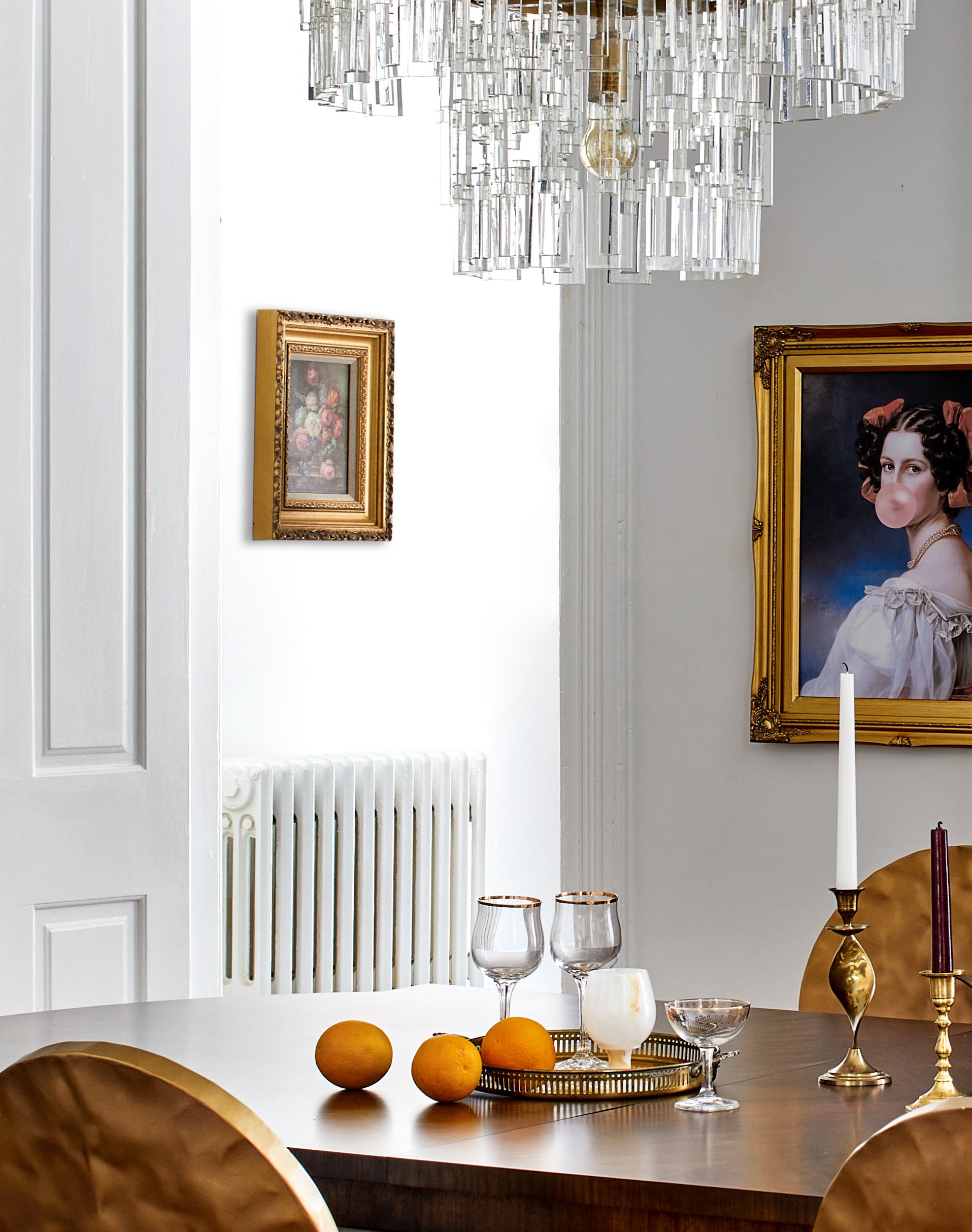Elegant dining room with a wooden table decorated with oranges, glassware, candlesticks, and a chandelier, with framed artwork on white walls.