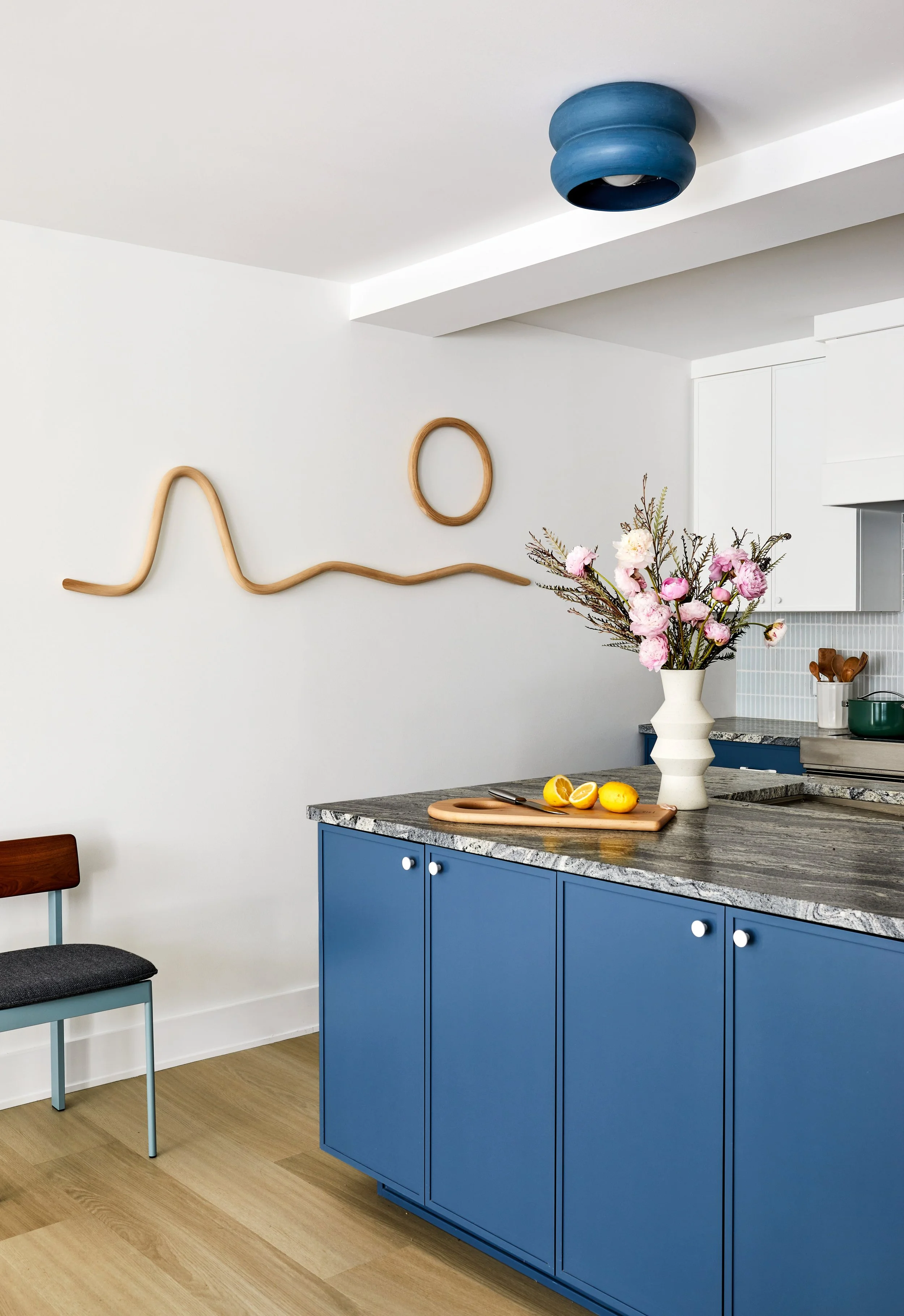 Kitchen with blue cabinets, granite countertop, white walls, pink flowers in a white vase, lemons on a cutting board, and wall art made of curved wooden sticks and a round loop.