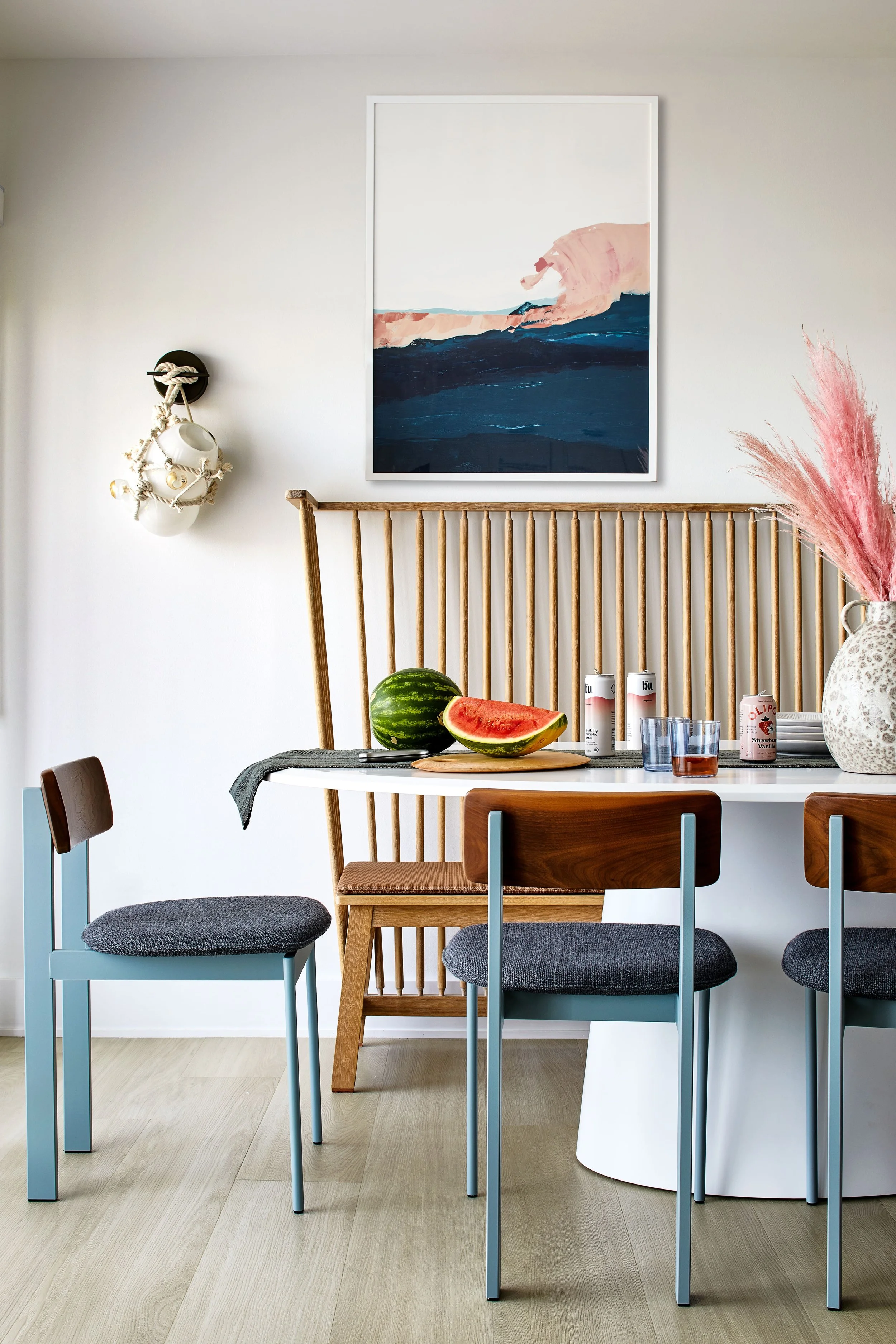 Modern dining area with a white table, three chairs, and a wooden bench. The table has a watermelon with a slice cut out, three cans of drink, and glasses. A vase with pink pampas grass decorates the table. A large abstract art piece depicting waves 