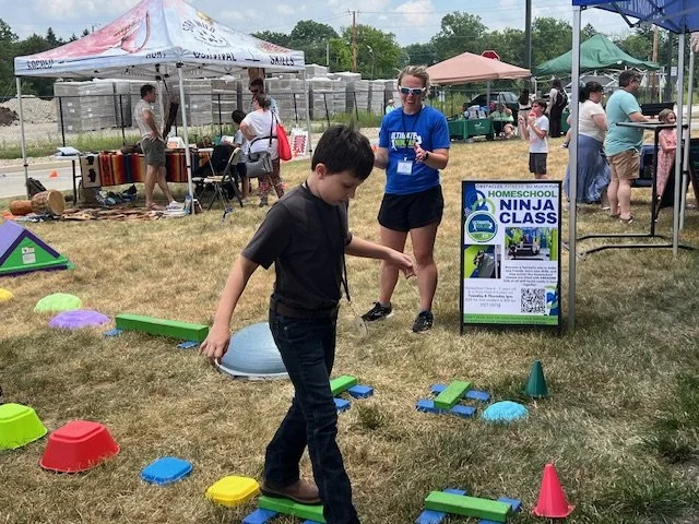 Boy balancing on a pedal plane at a homeschooling event with tents and people in the background.