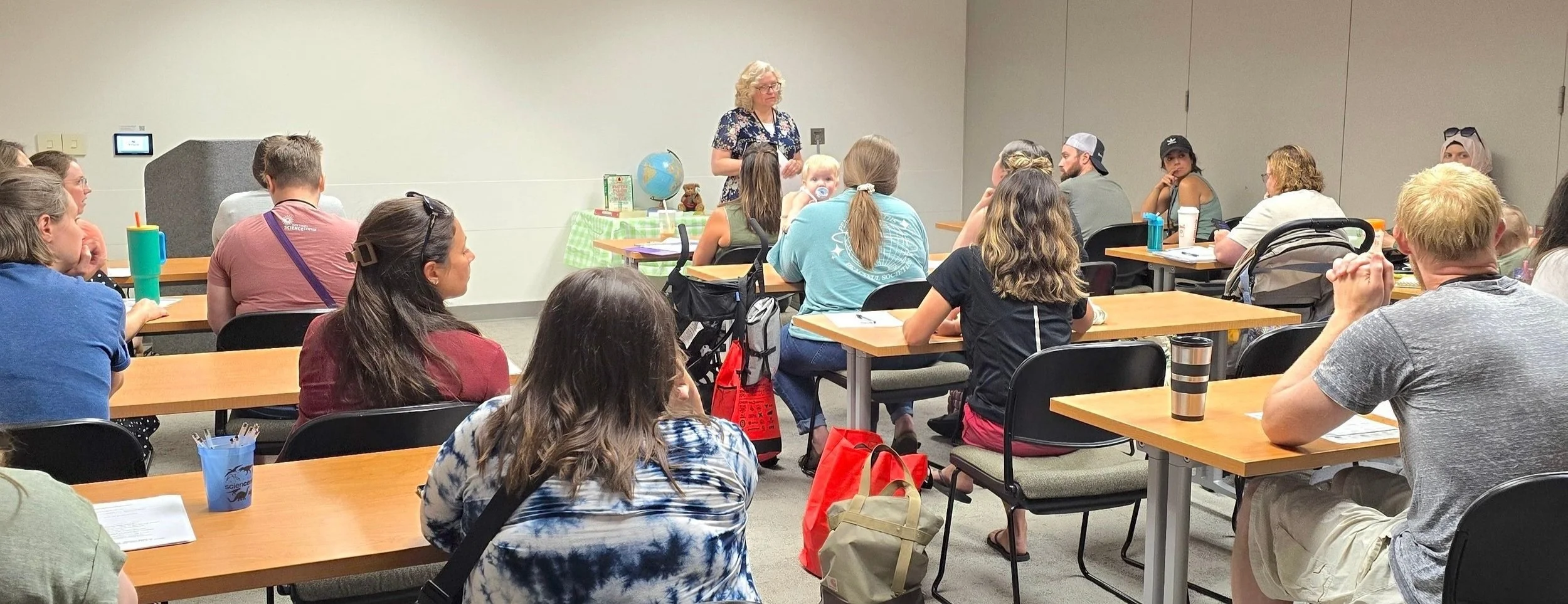 Instructor leading a presentation in a classroom with adult students seated at tables, some taking notes, some listening attentively, with educational materials and a globe on a table near the instructor.