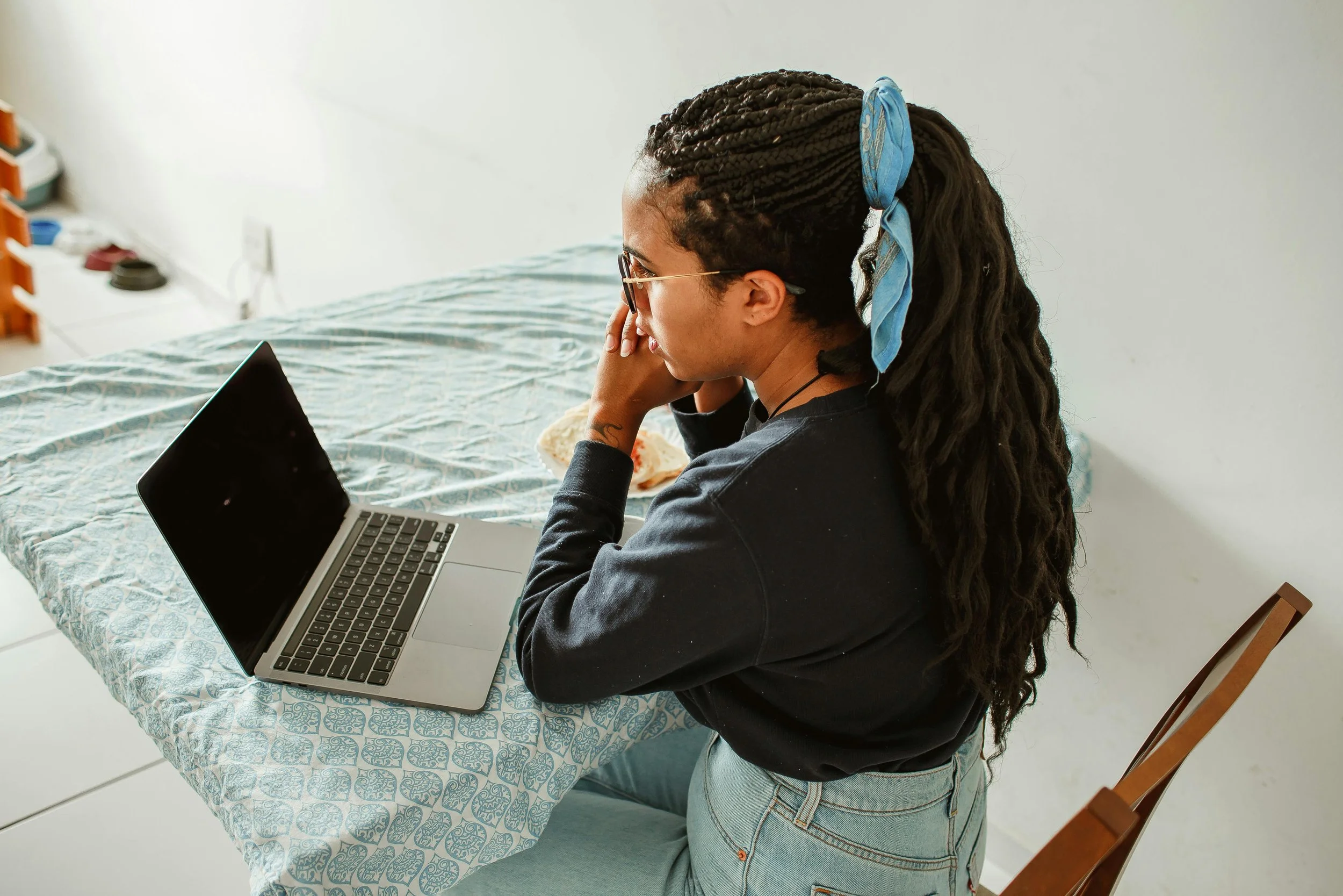 A young woman with braided hair and glasses sitting at a table, working on a laptop with a patterned tablecloth.