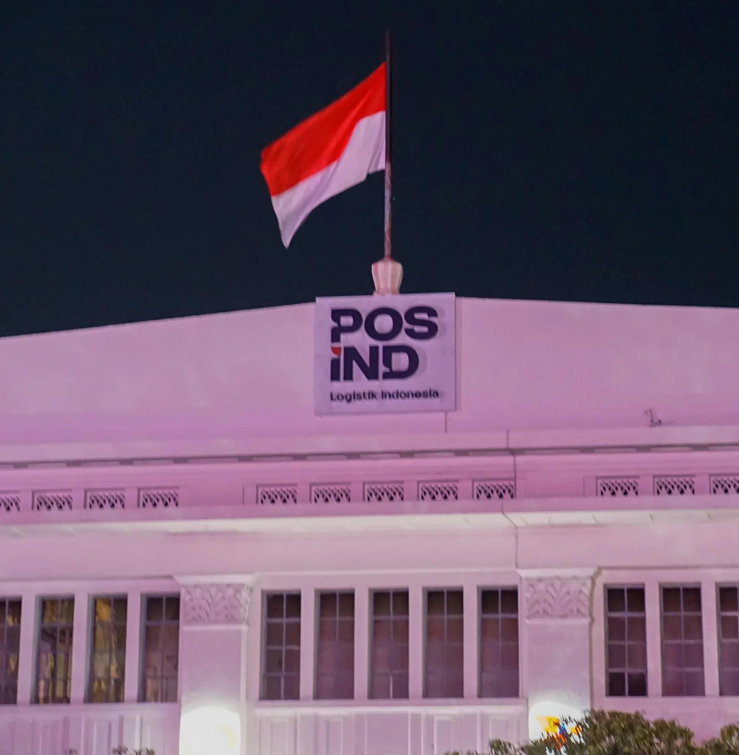 An Indonesian flag on a flagpole flying above a building with a sign that reads 'POS IND Logistics Indonesia' at night.