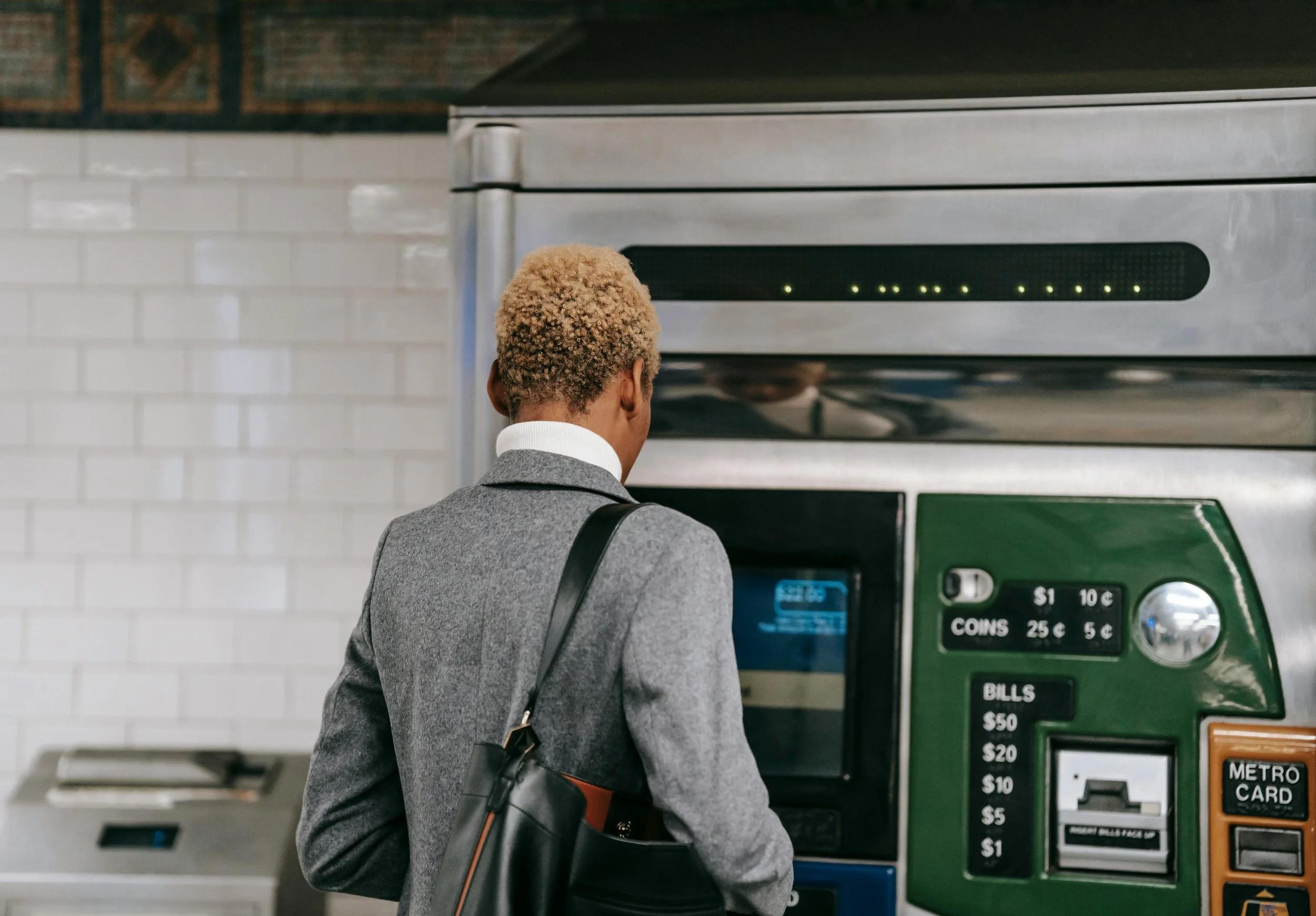 A person with blonde hair in a gray suit is using a ticket vending machine at a metro station.
