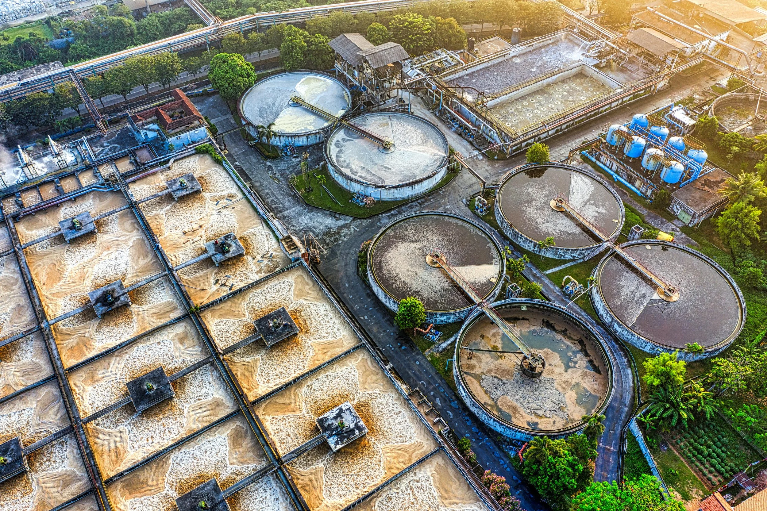 Aerial view of a water treatment plant with multiple large circular and rectangular tanks, some with water in different stages of processing, surrounded by green trees and vegetation.