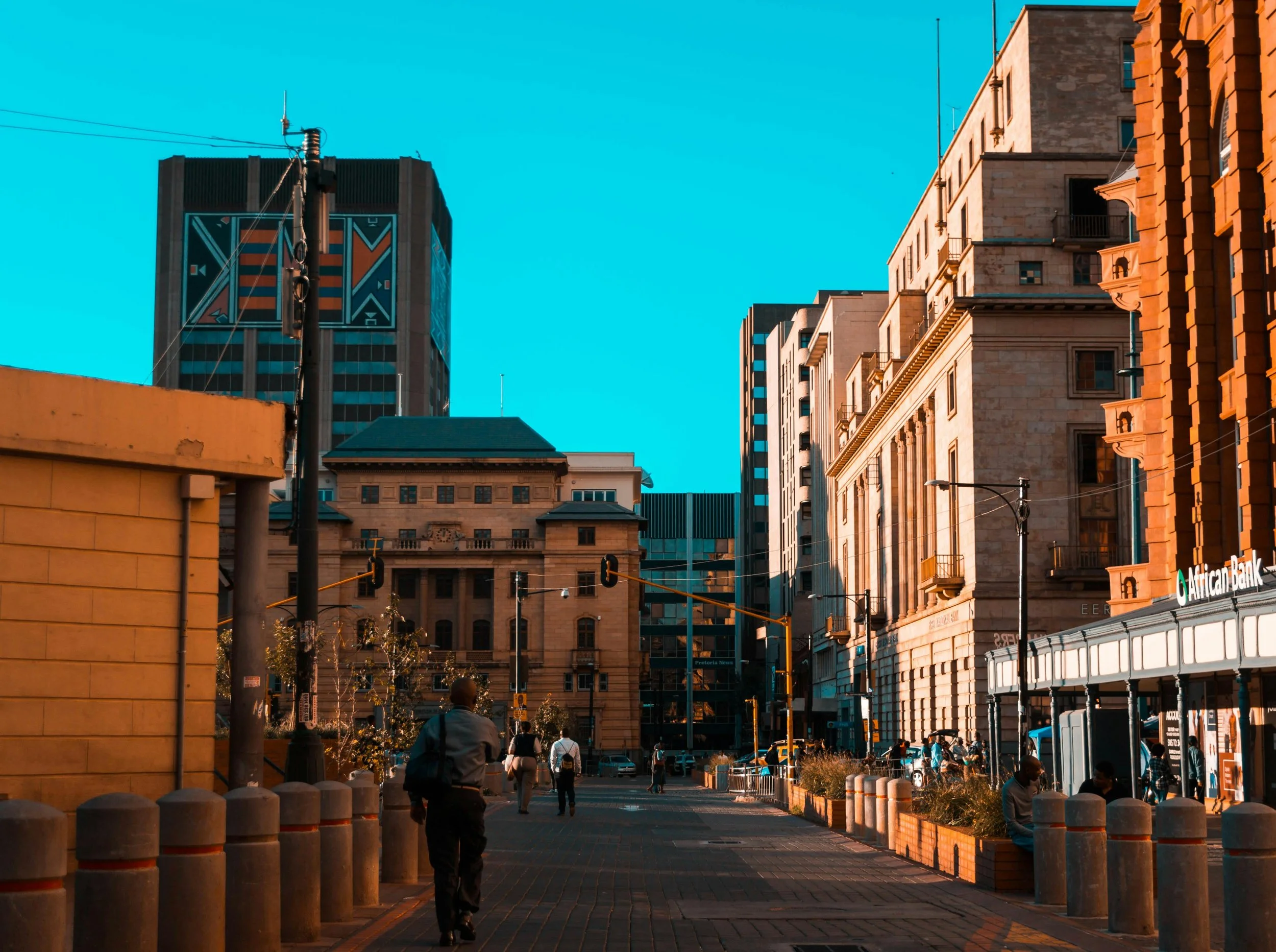 City street with tall buildings, some with modern glass facades and others with traditional architecture, under a clear blue sky. Pedestrians walking and streetlights visible.