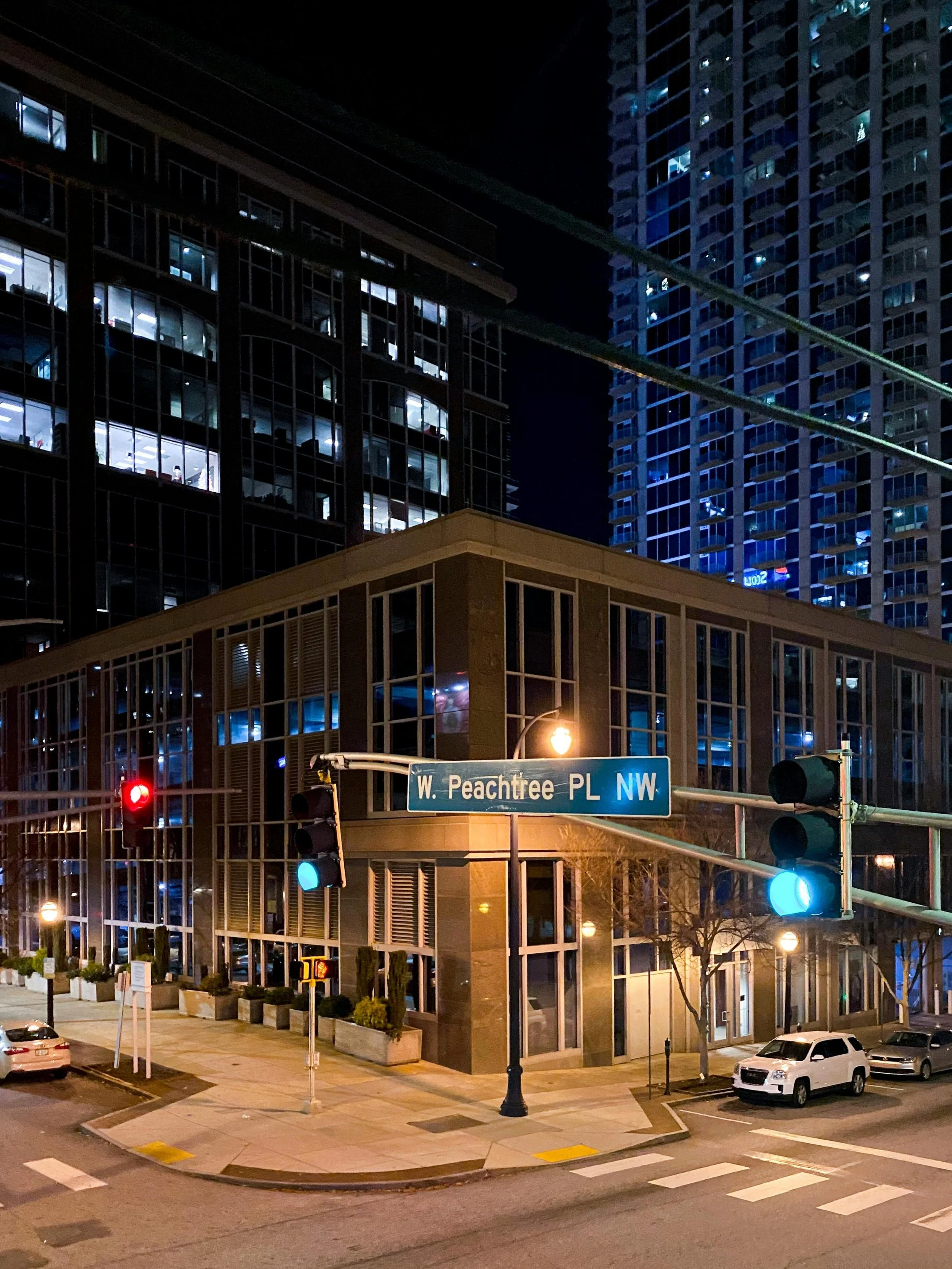 Nighttime city scene showing tall glass buildings with illuminated windows, traffic lights, street signs, parked cars, and sidewalk with trees and planters at the intersection of W. Peachtree Place NW.