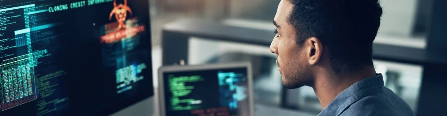 A man in a blue shirt working on multiple computer screens displaying code and cybersecurity information in a tech office.
