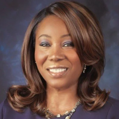 Portrait of a woman with shoulder-length light brown hair, wearing makeup, earrings, a necklace, and a navy blue top, smiling against a dark blue background.