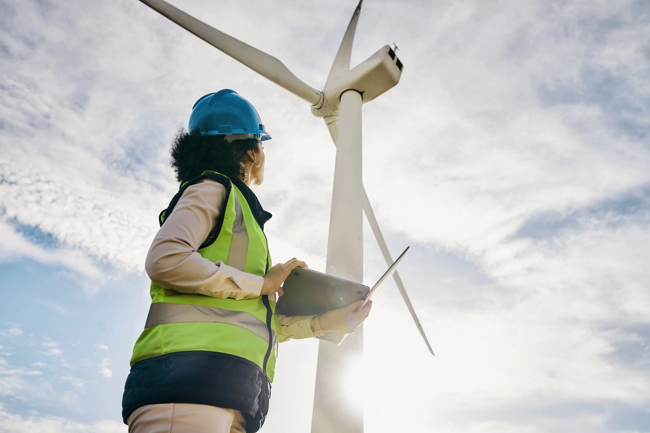 A woman wearing a blue safety helmet and a yellow safety vest inspecting a wind turbine with a tablet in her hand.