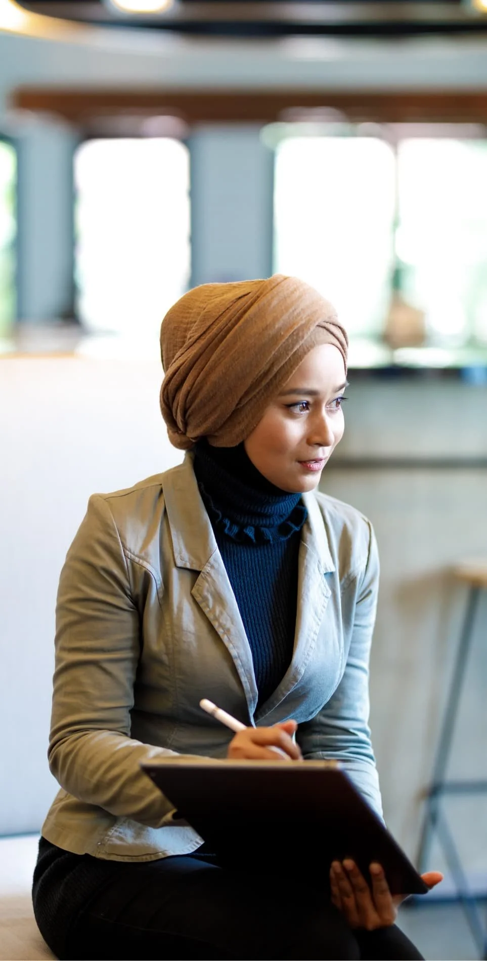 A woman wearing a brown headscarf and beige jacket, sitting and taking notes with a pen in a modern indoor space with large windows.