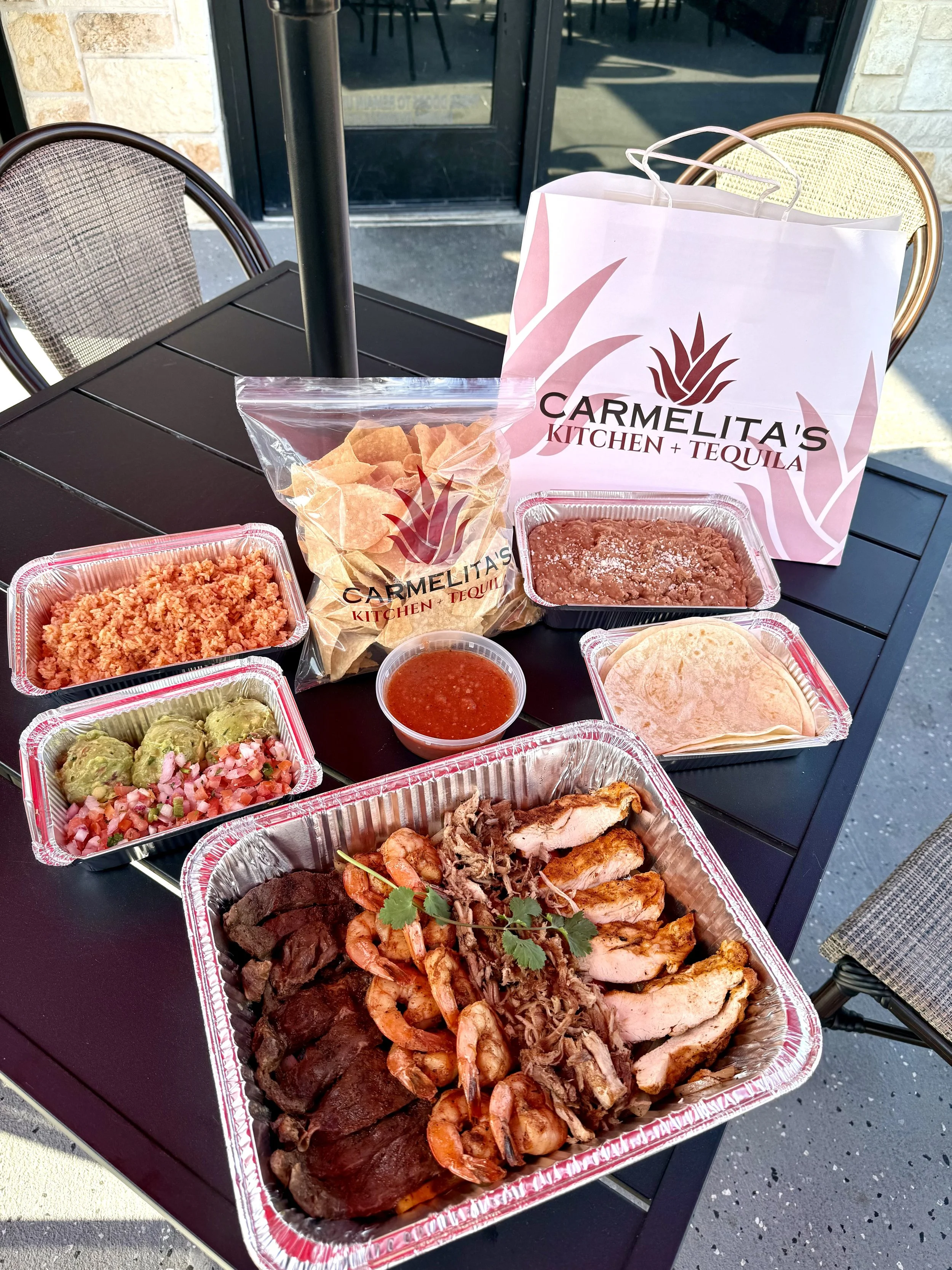 Outdoor table with Mexican food takeout from Carmelita's Kitchen + Tequila, including grilled chicken, shredded beef, shrimp, rice, guacamole, salsa, tortillas, and nachos, with Carmelita's shopping bag in background.