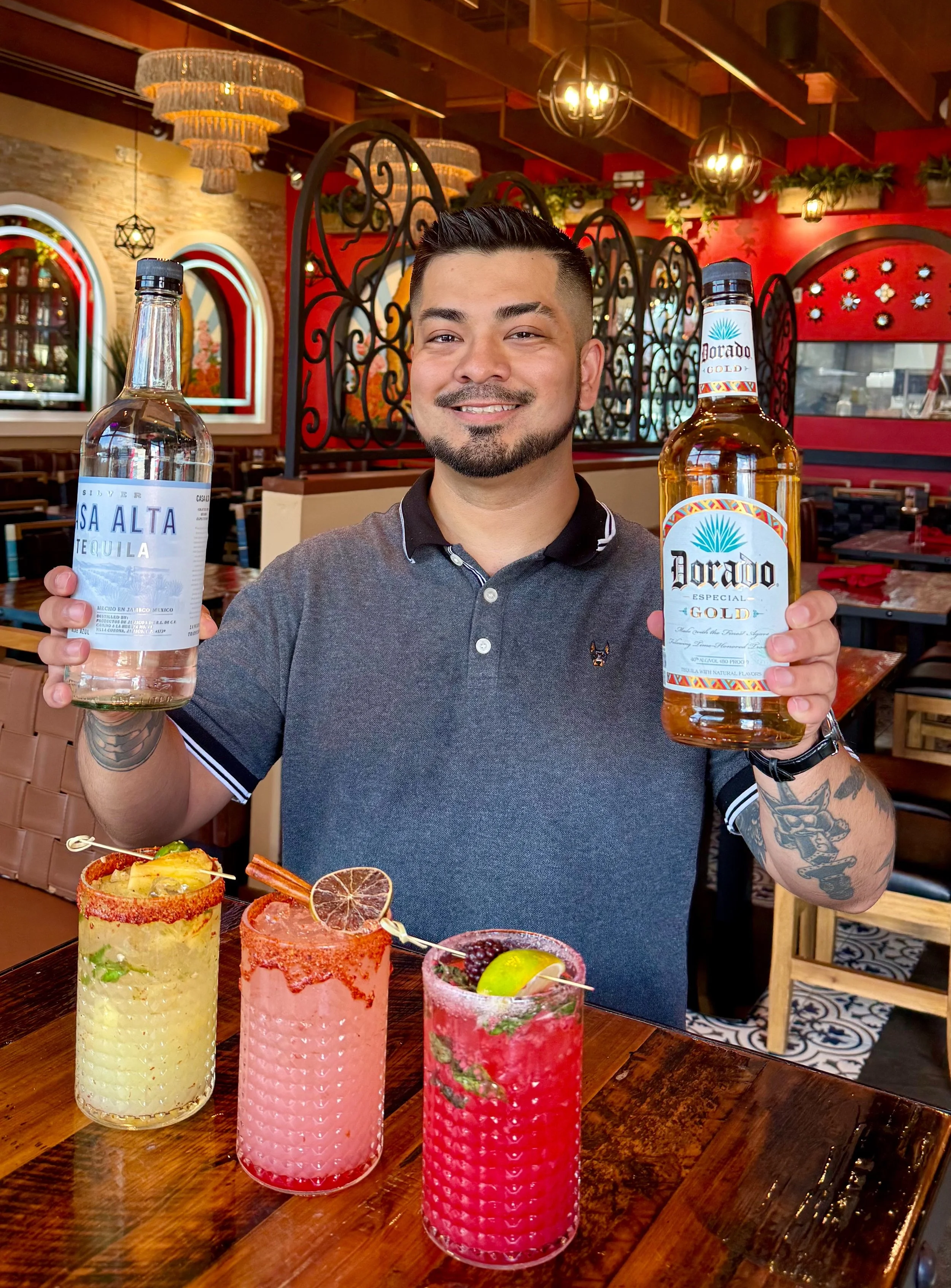 A man smiling and holding a bottle of silver Alta tequila in one hand and a bottle of Dorado Gold tequila in the other, seated at a table with three colorful cocktails garnished with fruits.