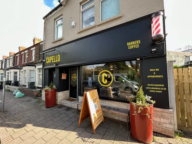 The exterior of a barbershop called Capello Barbers, with a black storefront featuring yellow signage. There are two large red and white barber pole lights on each side of the sign. In front, there are large red planters with small plants, a wooden stand with a chalkboard menu, and a QR code sign. The building is on a street lined with similar row houses.