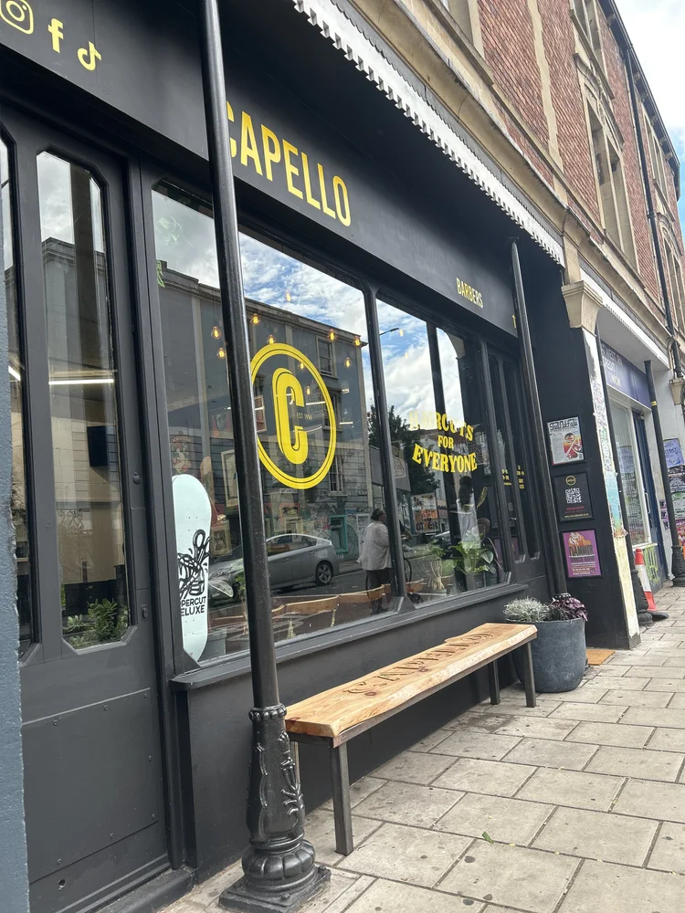 View of a black storefront with large glass windows and a black awning with yellow lettering that says 'GAPELLO' and 'BARBERS.' Inside the store, reflections of the street and sky are visible in the windows. Outside, there is a wooden bench, a black metal lamp post, and a large planter with flowers on a sidewalk.
