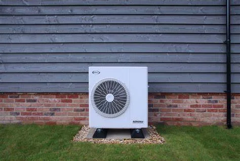 An outdoor air conditioning unit placed on a gravel pad against a brick and wooden siding wall.