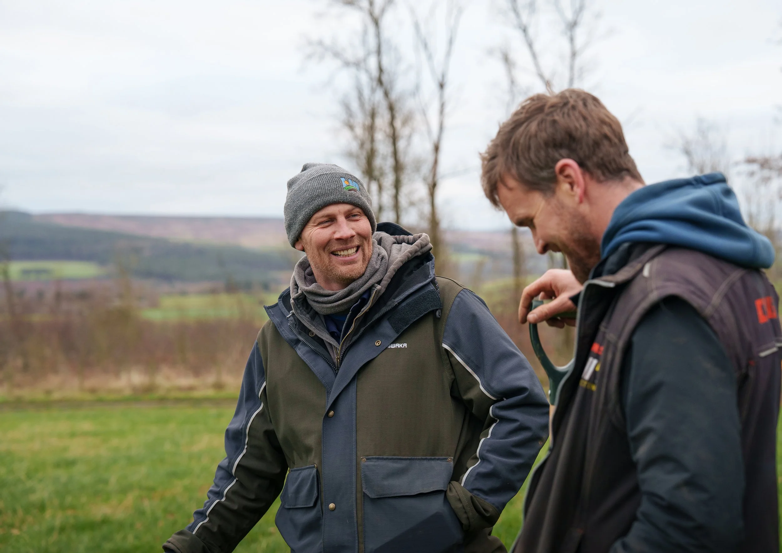 Two men outdoors on a cloudy day, standing in a grassy field with trees and hills in the background. One man is smiling and looking at the other, wearing a beanie, a scarf, and a jacket. The other man, wearing a hoodie and jacket, is holding a small object, possibly a camera or binoculars, and is looking down at it.