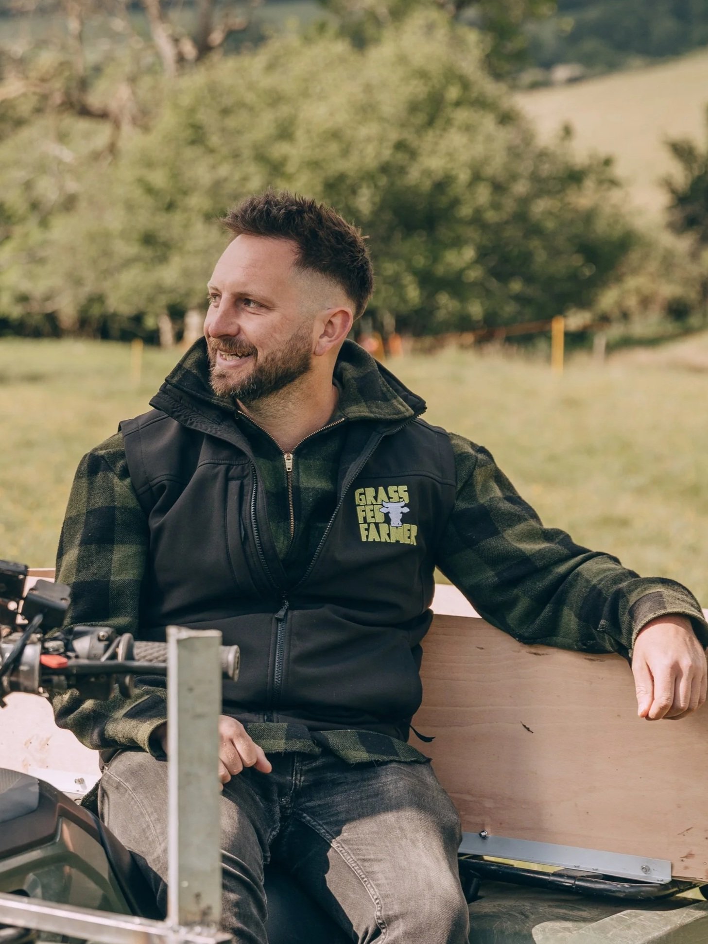 A man with a beard and short dark hair sitting on a bench outdoors, wearing a black vest with a logo that reads 'GAS FARMER' over a green camouflage shirt, smiling and looking to the side with a blurred green landscape in the background.