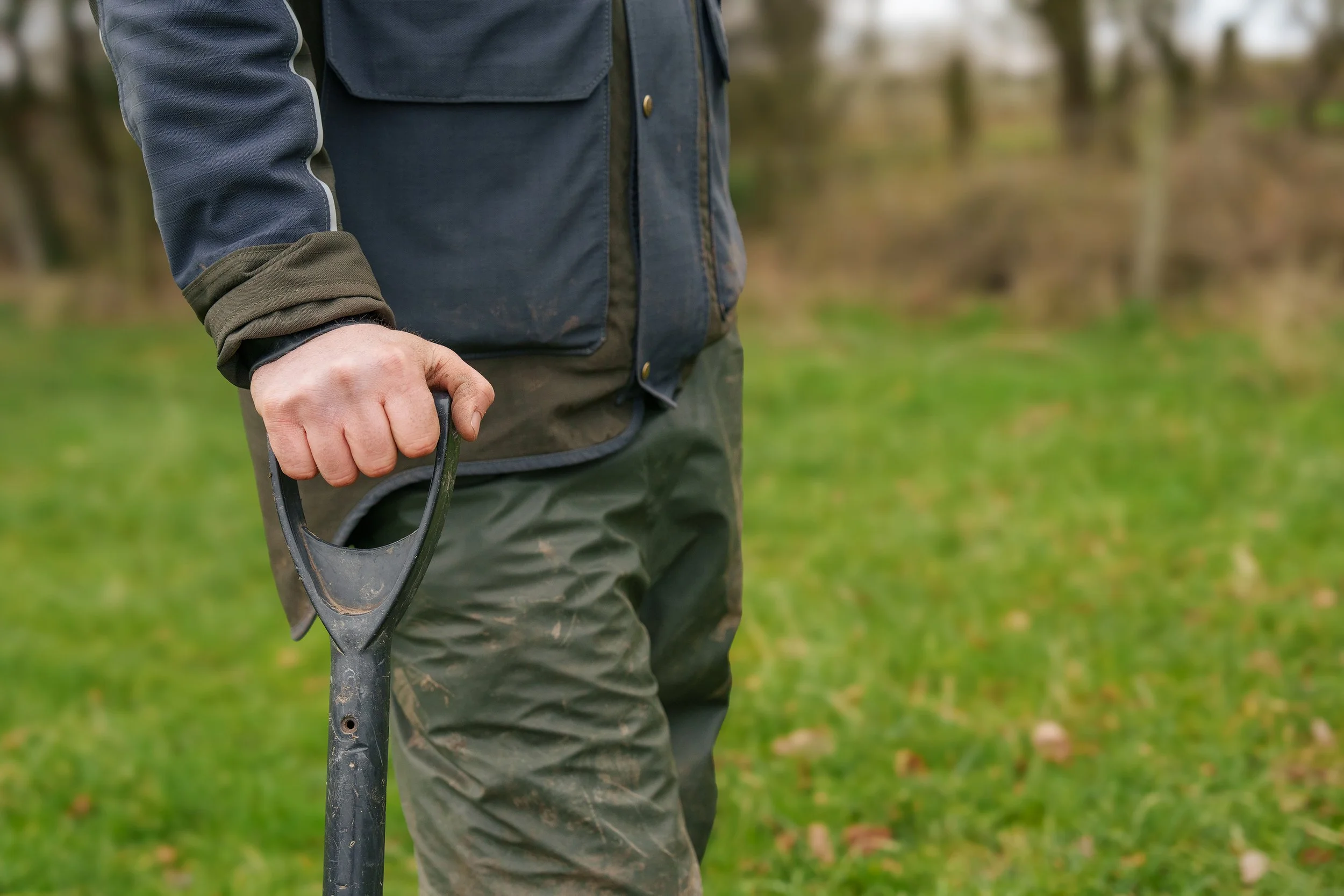 Close-up of a person wearing a dark jacket and green pants, holding a black shovel handle outdoors on grass with blurred trees in the background