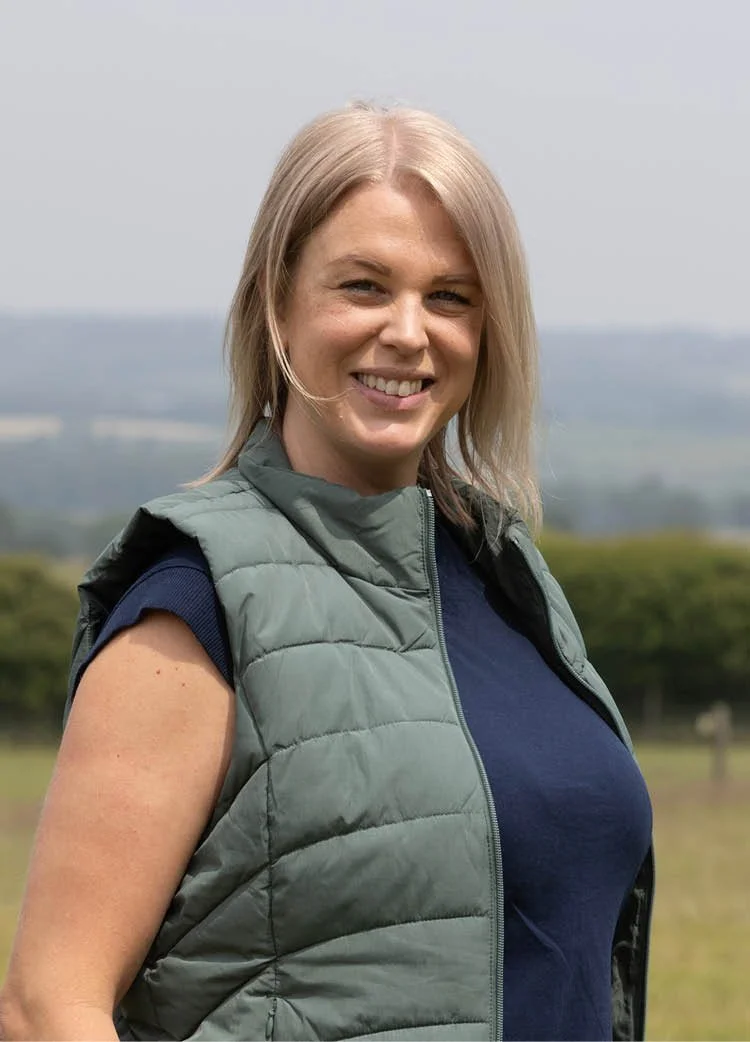 A woman with blonde hair smiling outdoors in a blue shirt, with a background of trees and grass.