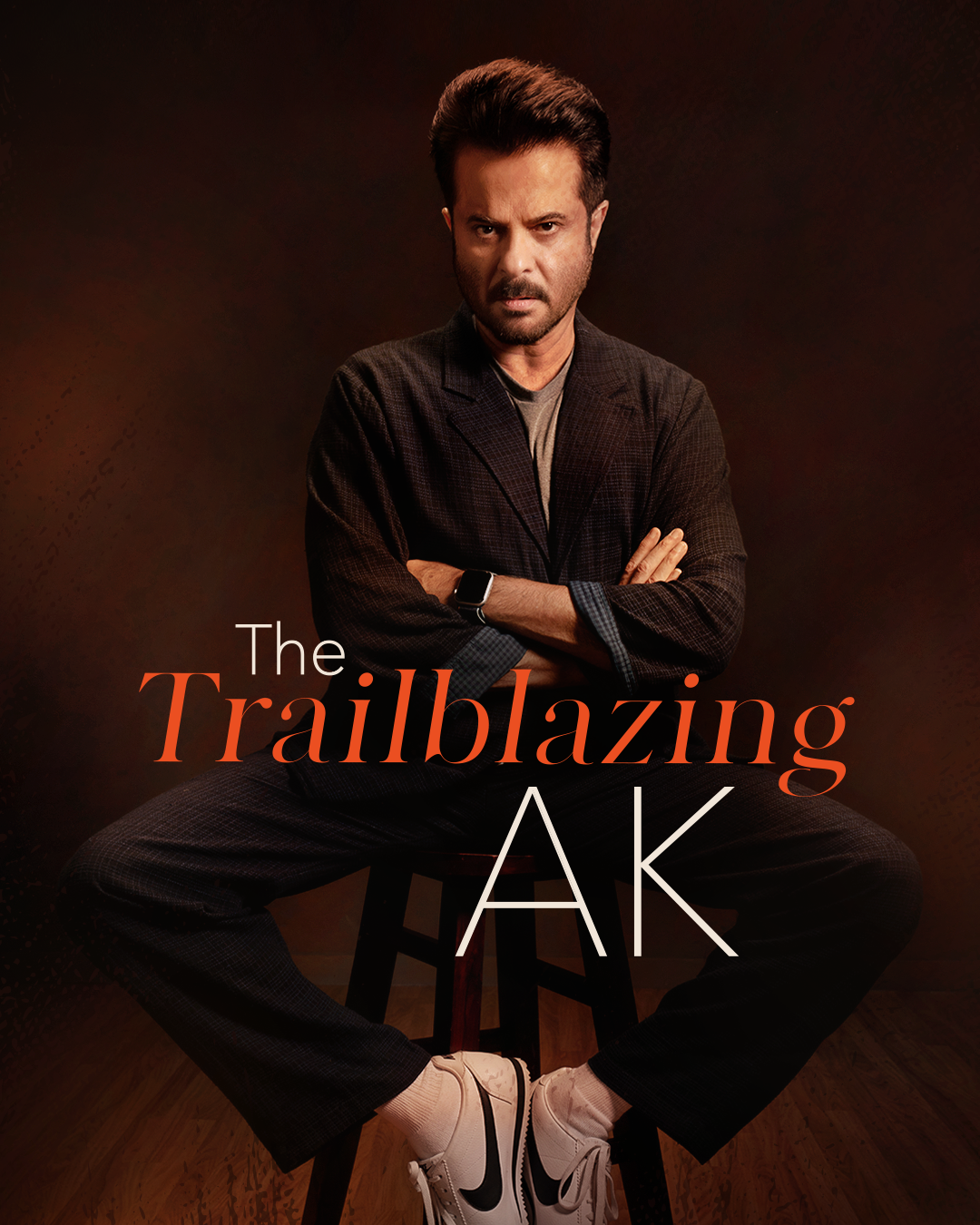 A person sitting confidently on a stool with arms crossed, wearing a dark suit, gray shirt, and white sneakers. Text reads 'The Trailblazing AK' against a dark, textured background.