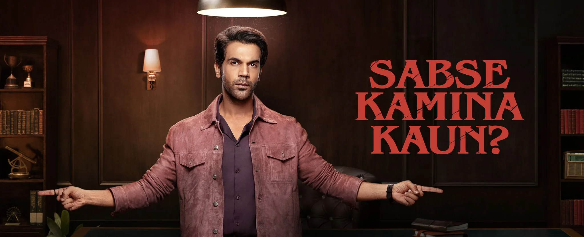 Man in a jacket with arms outstretched in a dimly lit room with bookshelves and trophies, text reads "SABSE KAMINA KAUN?" on the right.