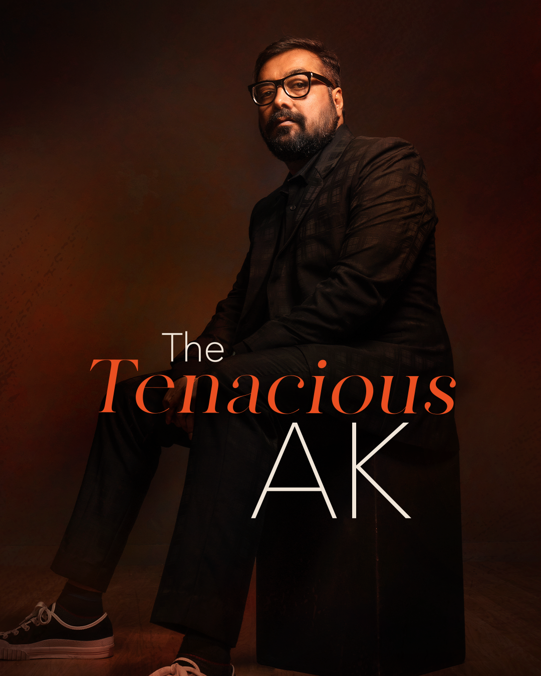 Man with glasses in a dark suit sitting on a stool, with 'The Tenacious AK' written in bold letters.