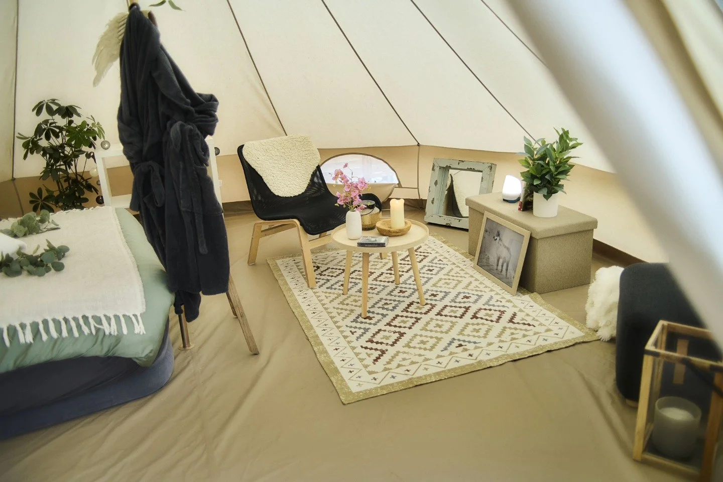 Intérieur d'une tente aménagée avec un tapis, une petite table ronde, un fauteuil, un lit avec une couverture blanche, et divers objets décoratifs et plantes