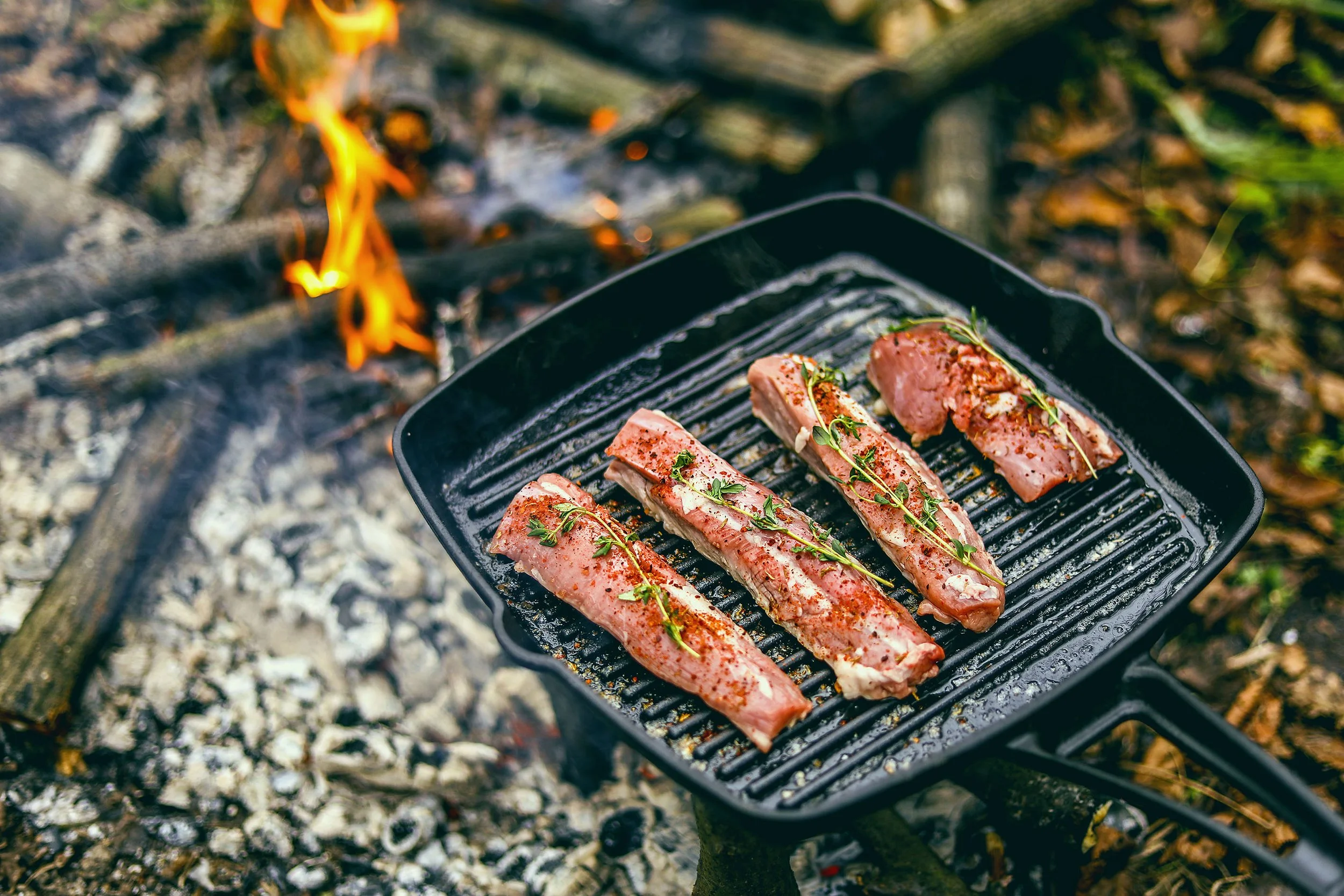 Des tranches de viande en train de cuire sur un gril, garnies de petites herbes, avec un feu de camp en arrière-plan.