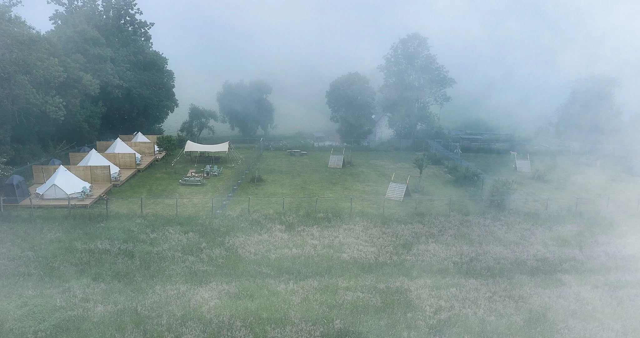 Campsites avec de petites tentes blanches et une grande tente, situés dans une zone herbeuse entourée d'arbres, avec un peu de brouillard.
