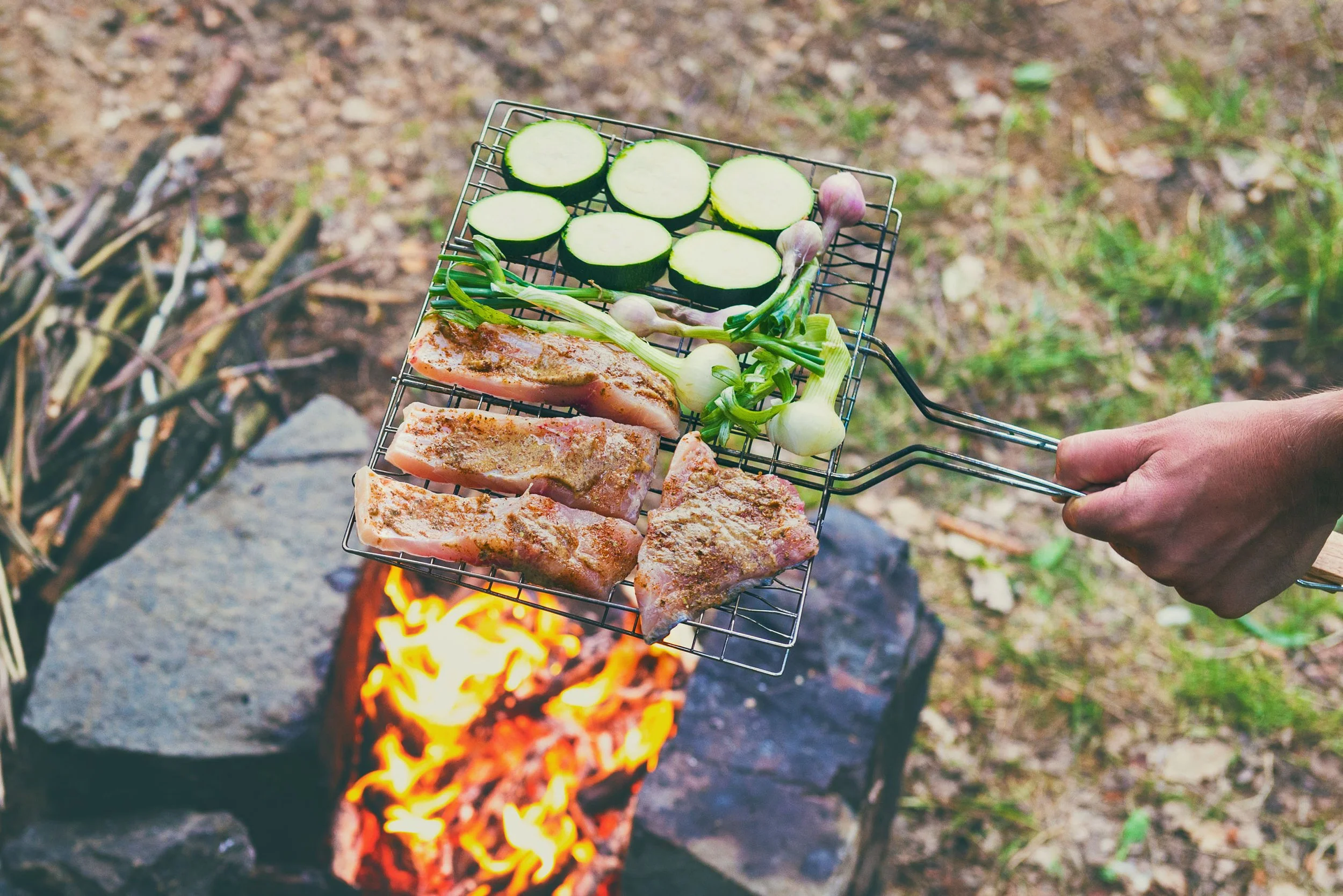 Une grille de cuisson avec des côtelettes de porc, des courgettes coupées en rondelles et des oignons verts, cuisine au-dessus d'un feu de bois en plein air.