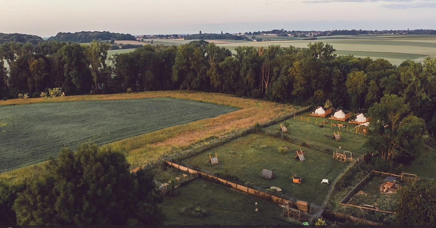 Champ en plein air avec quelques cabanes, un terrain de jeu pour chiens, entouré d'arbres et de champs agricoles au loin.