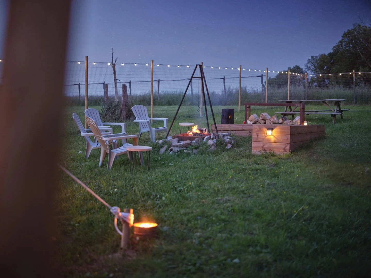 Zone de camping en plein air avec un feu de camp, des chaises en plastique, une table en bois, un panier de pierres, une clôture en filet avec des lumières suspendues et une table de pique-nique en bois, dans un environnement naturel en soirée.