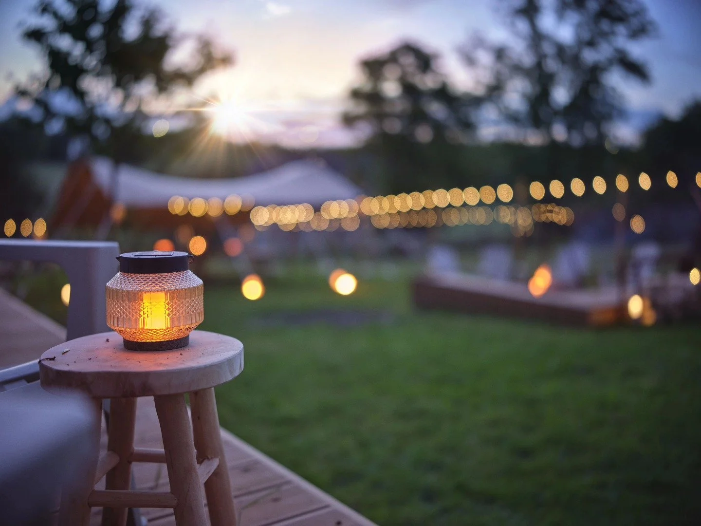 Une lanterne allumée posée sur un petit tabouret en bois, avec un fond flou de lumières décoratives en soirée dans un espace extérieur vert, avec des arbres et un ciel au coucher du soleil.