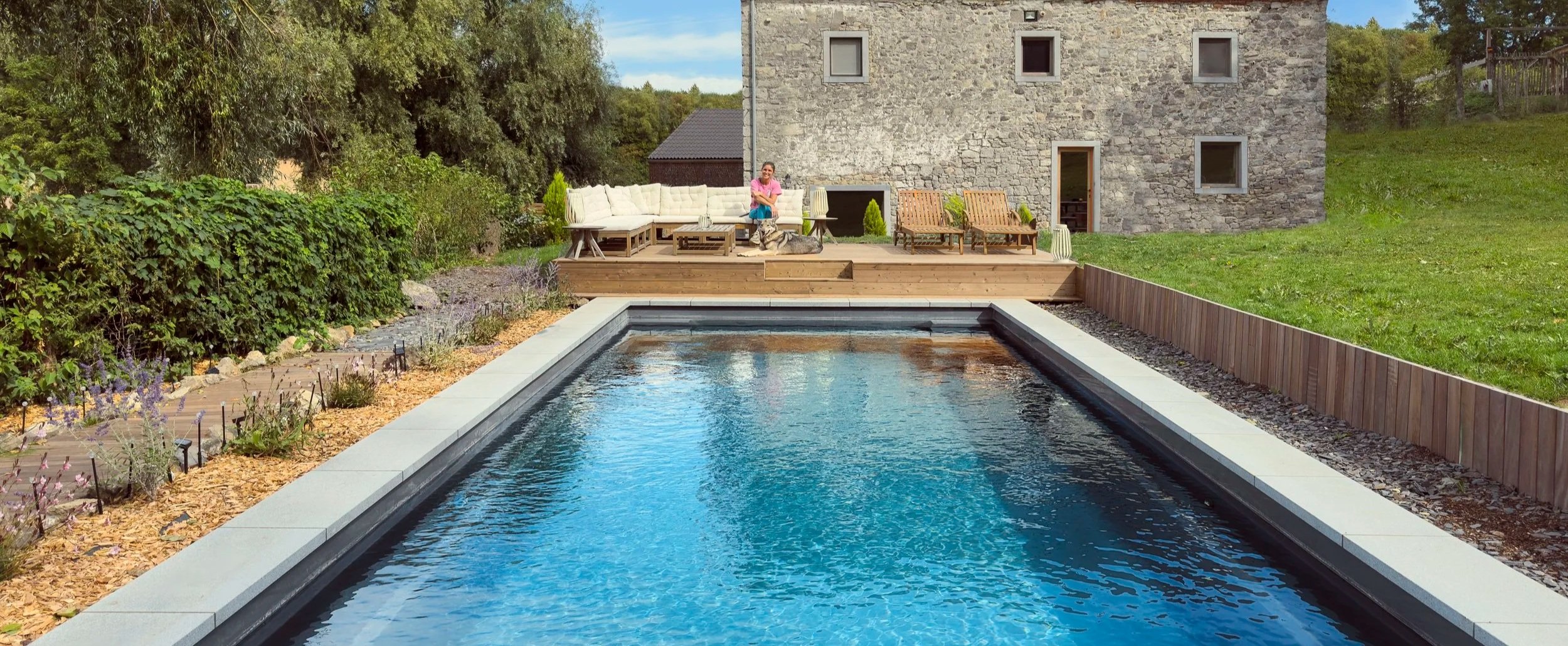 Une piscine dans un jardin entouré de végétation, avec un espace détente pourvue de mobilier en bois et une femme assise avec un chien, à l'arrière d'une maison en pierre.